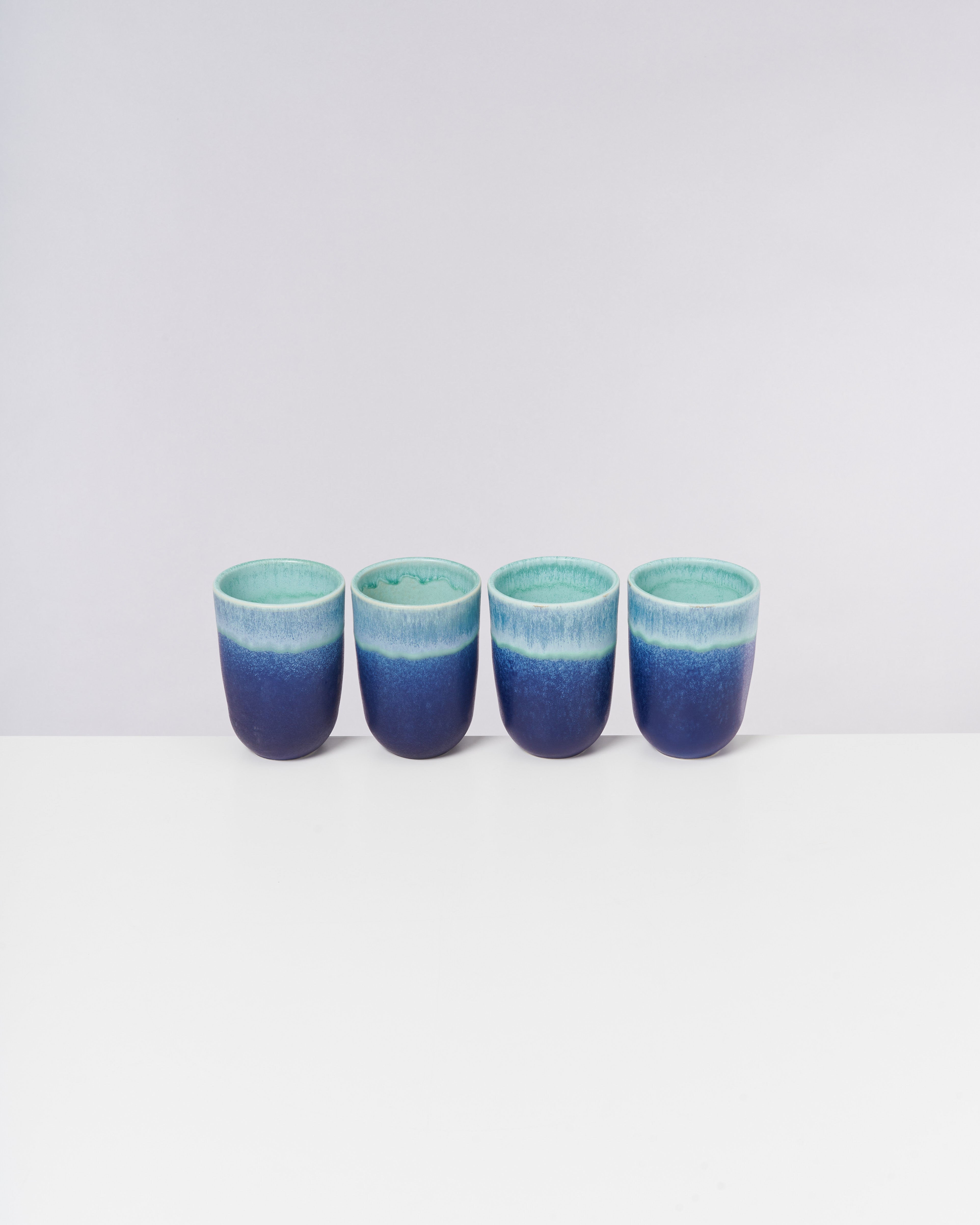 Vier blaue Becher mit einem türkisfarbenen Rand stehen nebeneinander auf einer weißen Oberfläche vor einem hellen Hintergrund.
