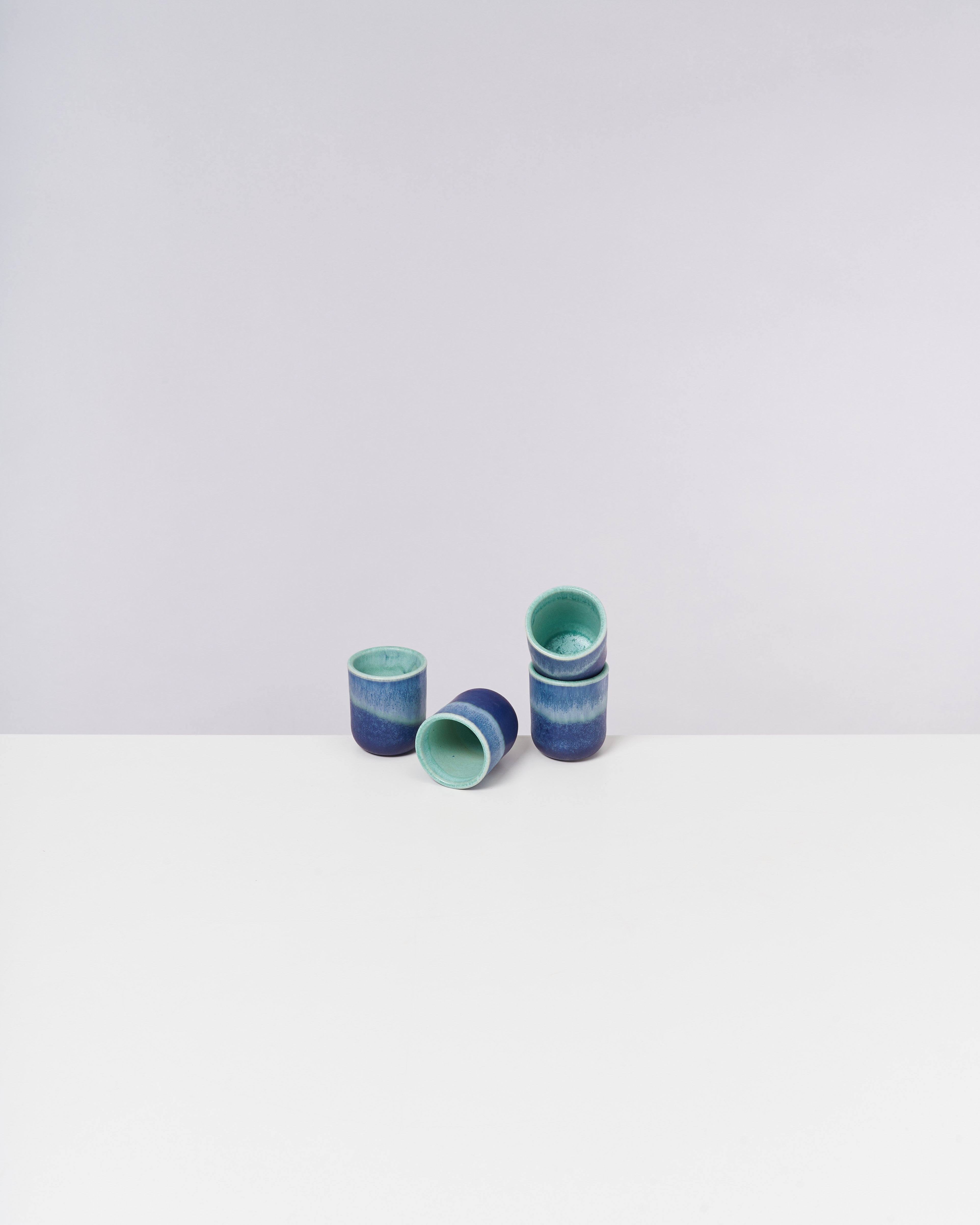Fünf Keramikbecher in Blau- und Grüntönen auf weißer Oberfläche vor grauem Hintergrund.