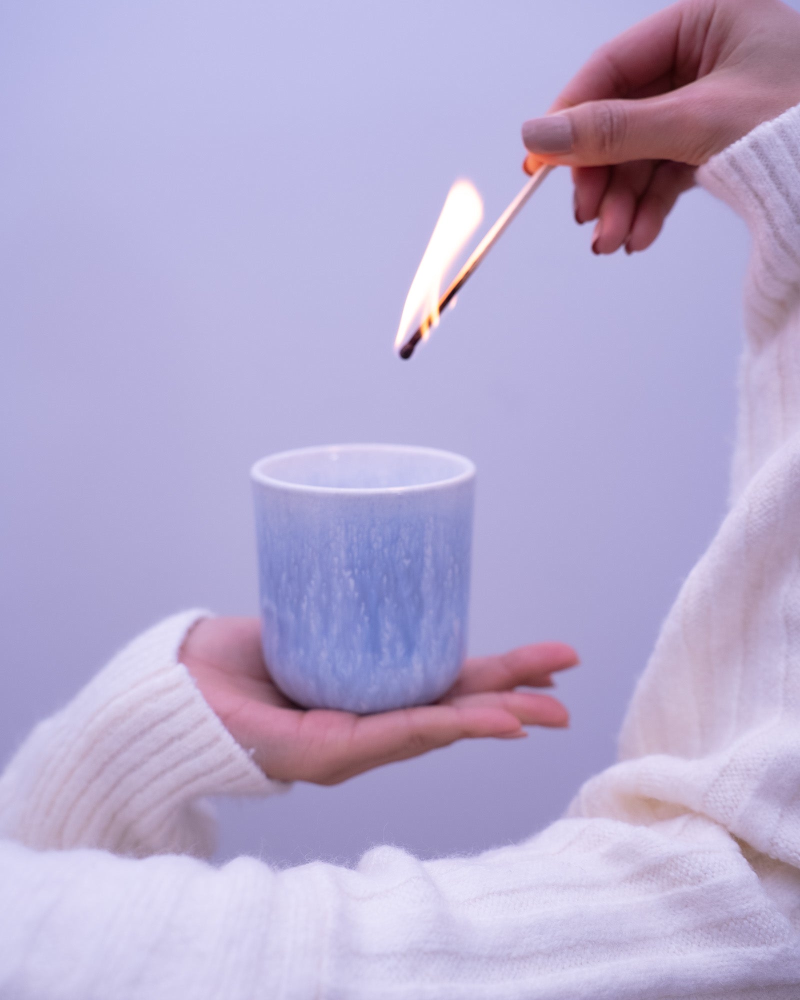 Hände in weißen Pullovern halten eine Kerze und zünden sie mit einem brennenden Streichholz an.