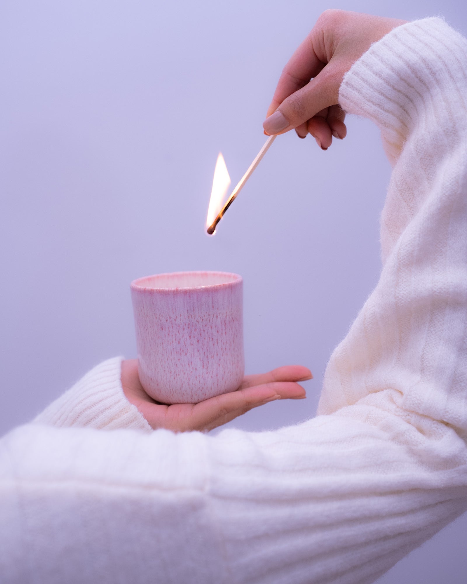 Eine Hand im weißen Pullover zündet mit einem Streichholz eine Kerze in einem rosa Becher an.