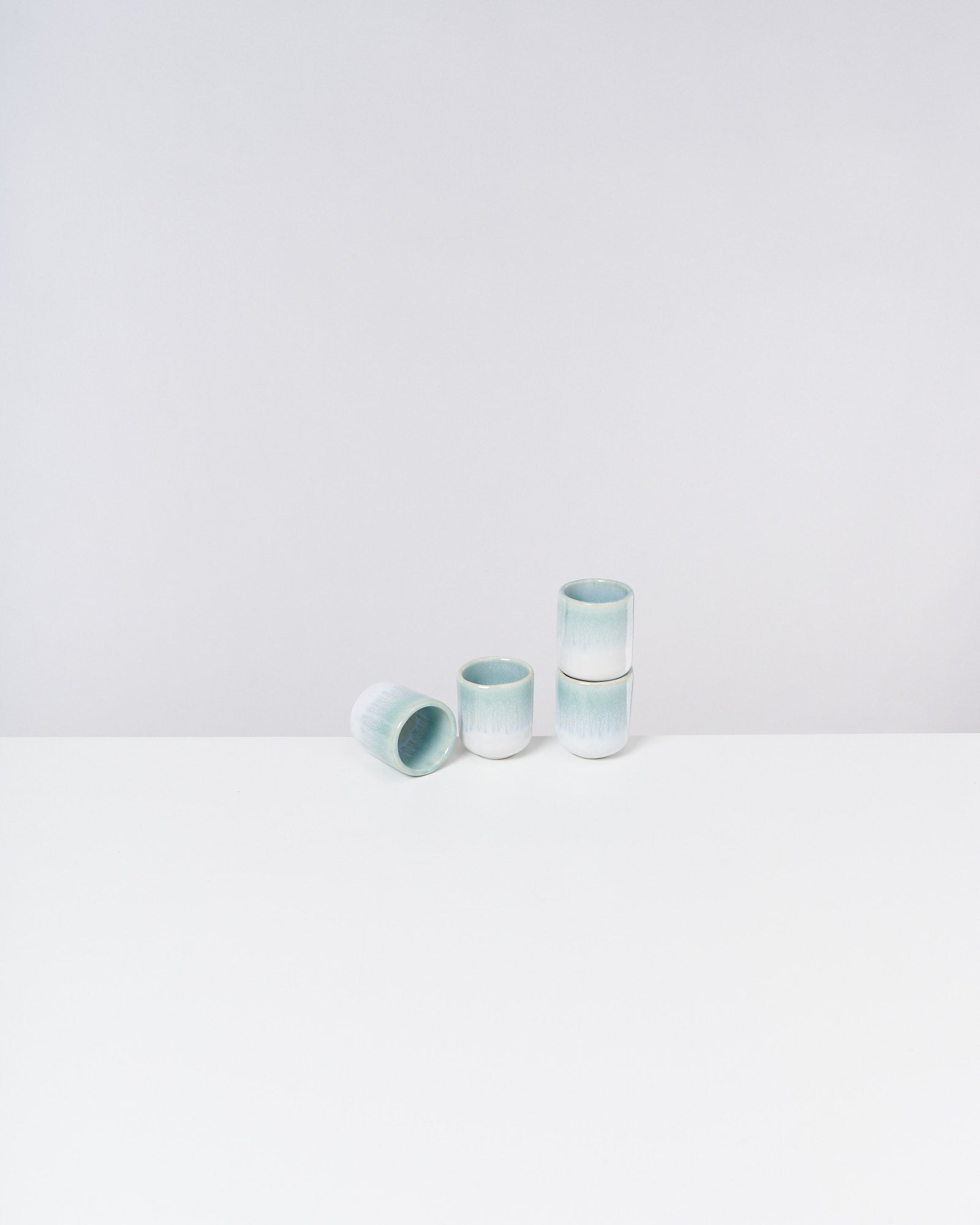 Drei türkisfarbene Becher auf einem weißen Tisch, einer liegt auf der Seite, zwei stehen gestapelt.