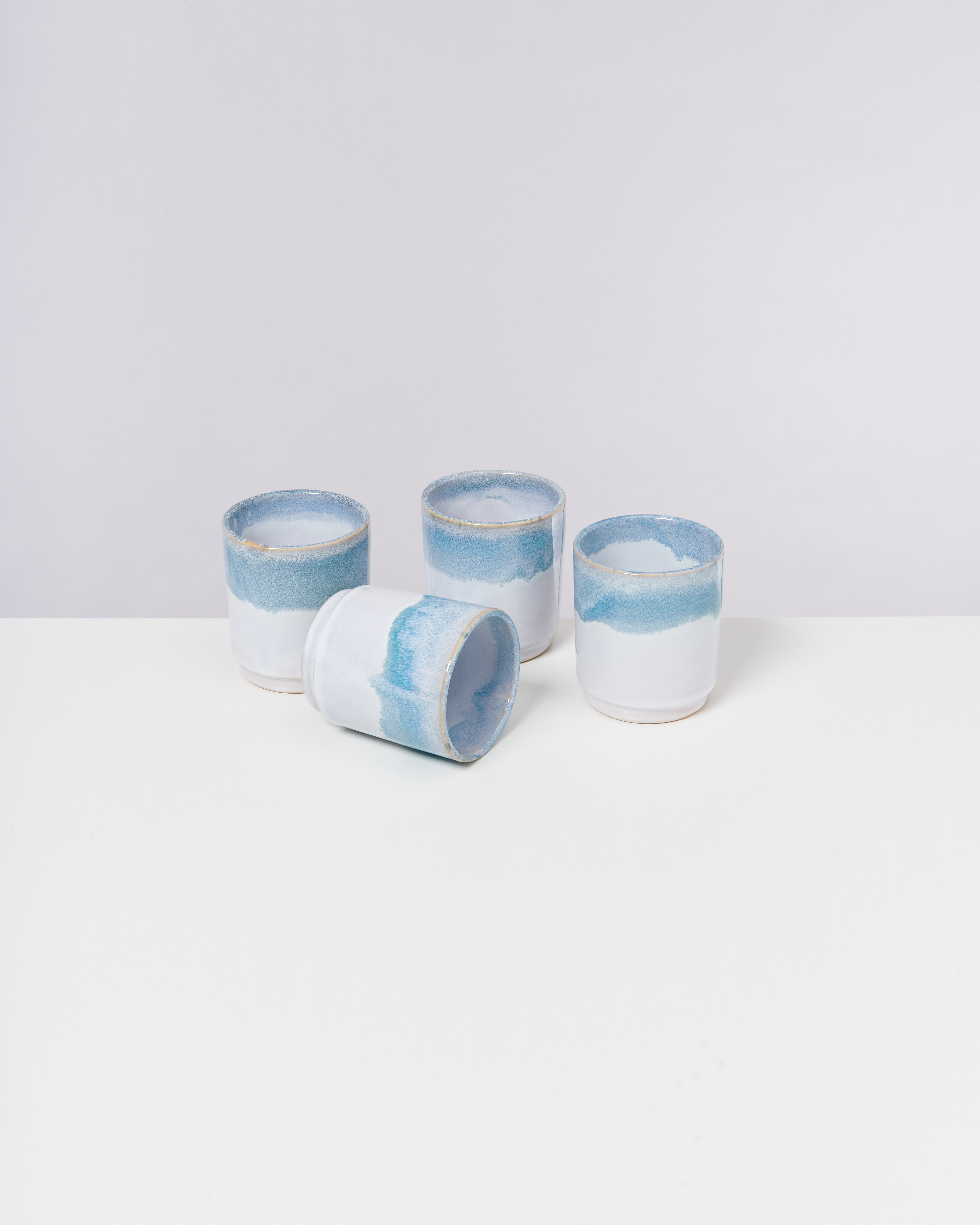Vier weiß-blaue Keramikbecher stehen auf einem weißen Tisch, einer liegt seitlich.