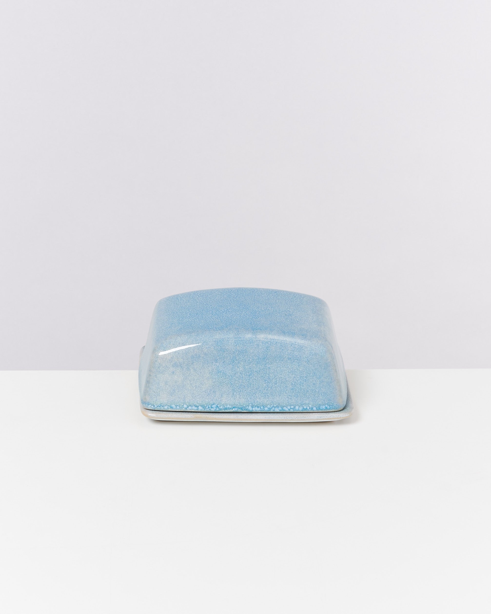 Blaue, rechteckige Keramikbutterdose mit Deckel auf einem weißen Tisch vor hellem Hintergrund.