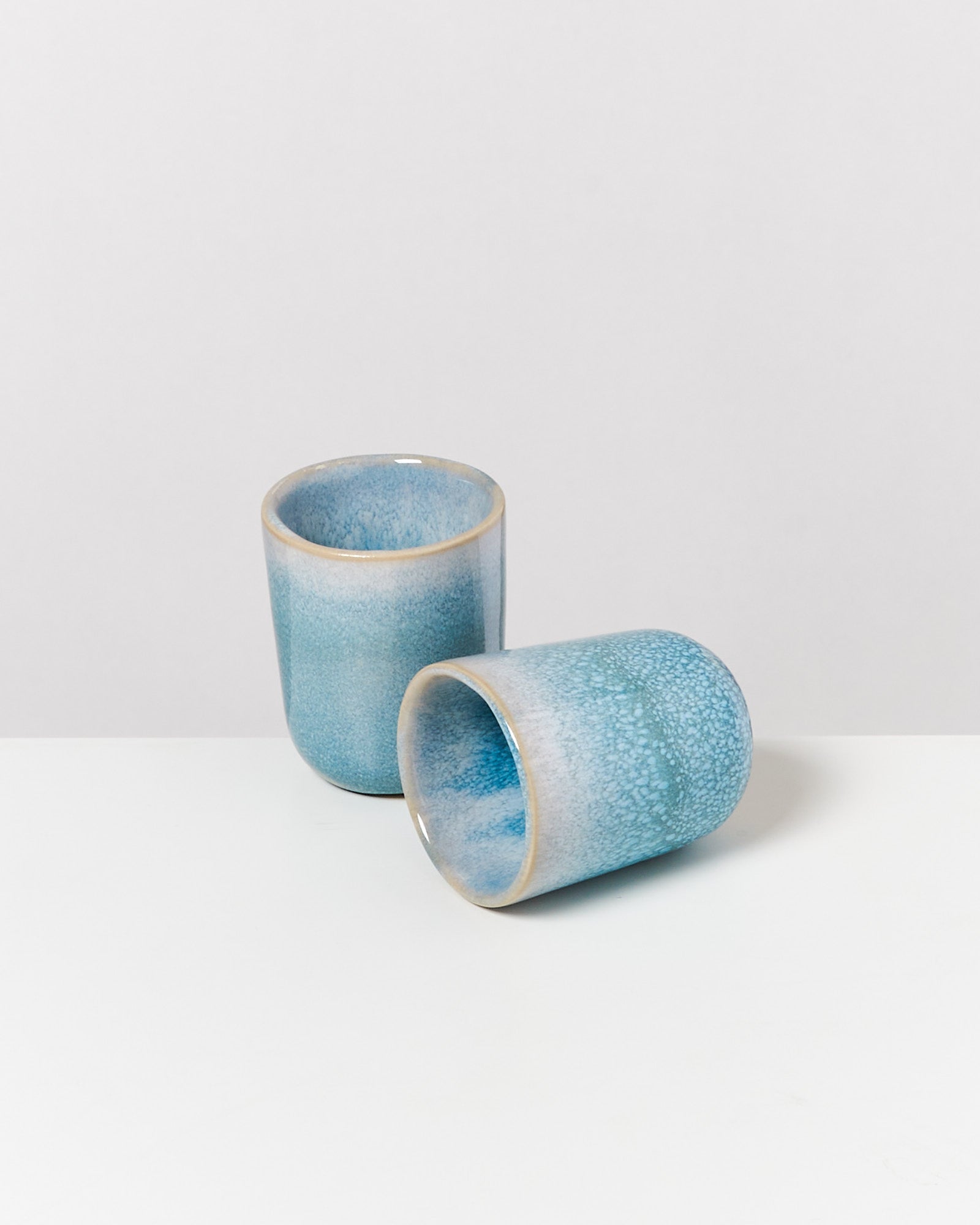 Zwei blaue Keramikbecher, einer stehend und einer liegend, auf einem weißen Hintergrund.