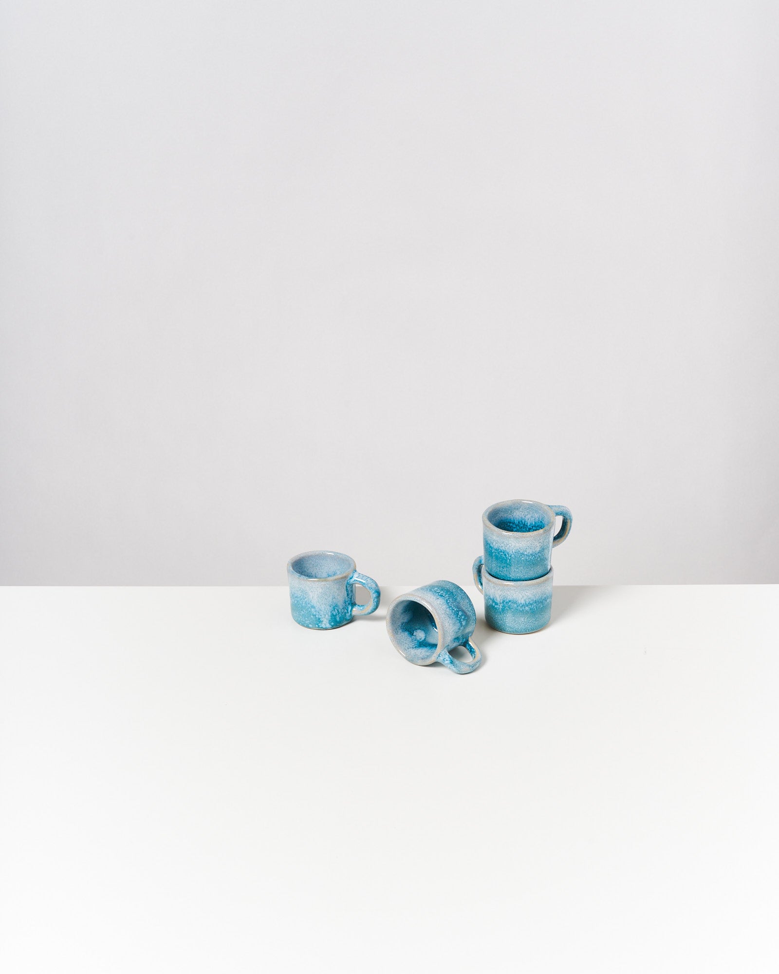 Drei türkisfarbene Keramiktassen auf einem weißen Tisch, eine liegt seitlich, die anderen sind gestapelt.