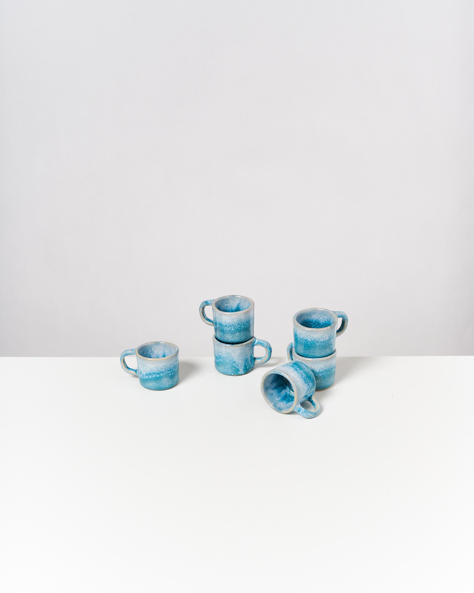 Sechs blaue Keramiktassen, teils gestapelt, stehen auf einem weißen Tisch vor einem neutralen Hintergrund.