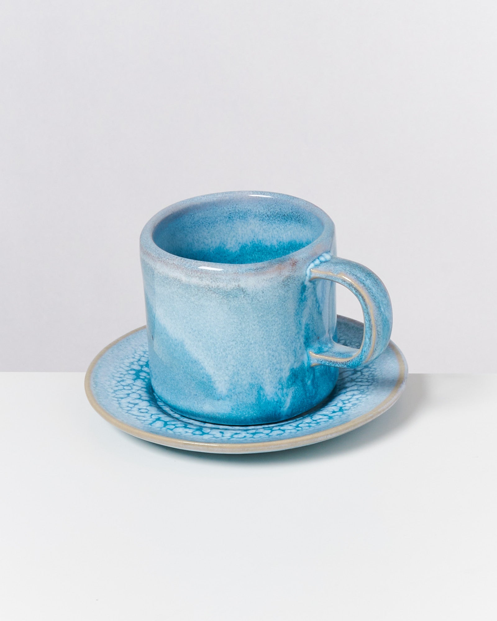 Blaue Tasse auf passender Untertasse mit marmorierter Glasur vor weißem Hintergrund.
