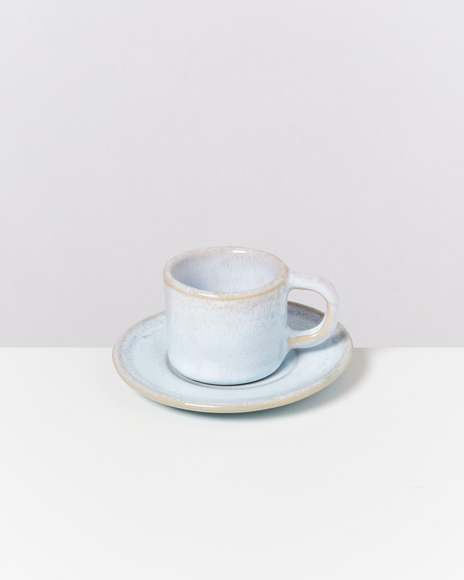 Blaue Keramiktasse auf einer passenden Untertasse, auf einem weißen Tisch vor einem hellgrauen Hintergrund.