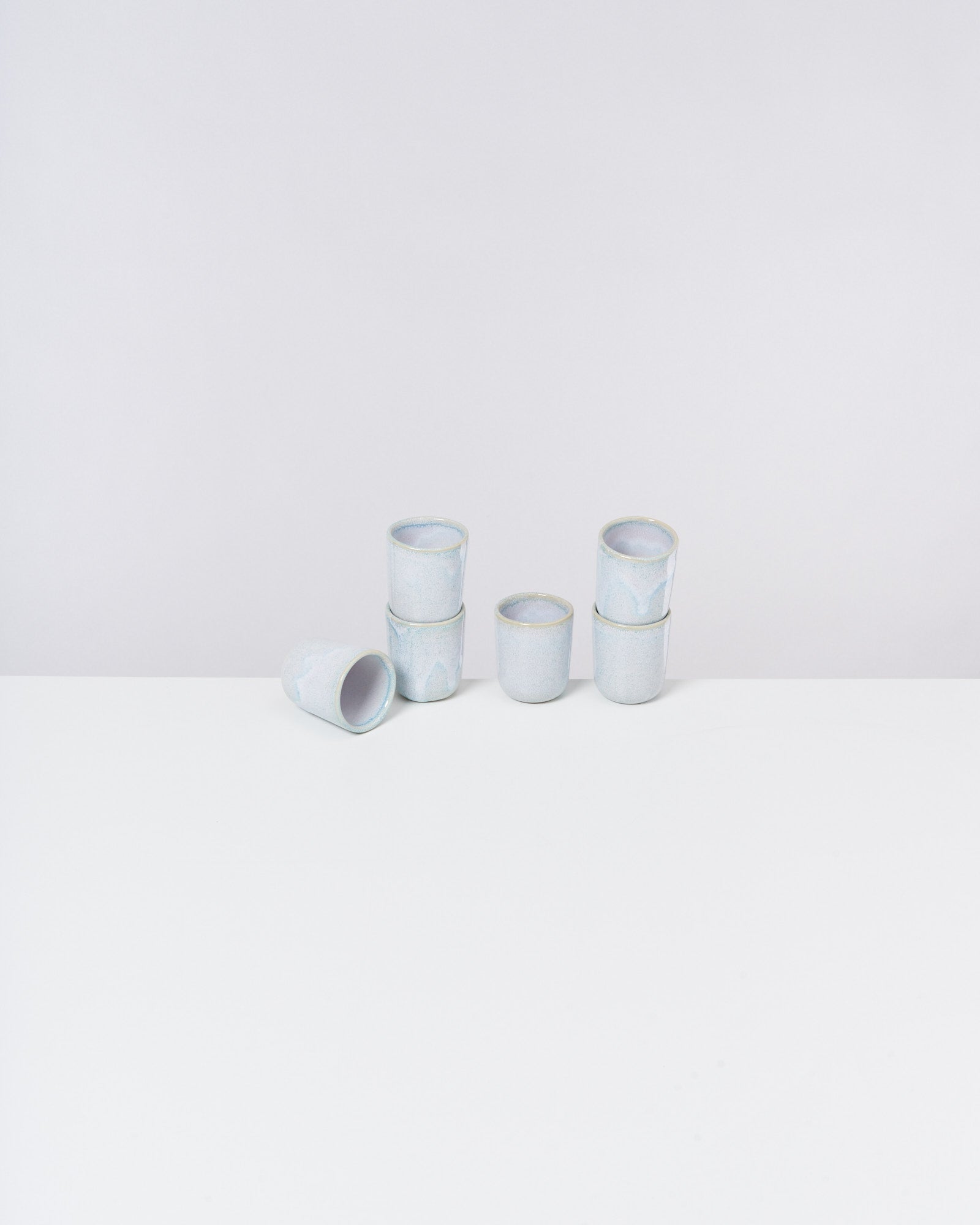 Sechs kleine, stapelbare Becher aus weißer Keramik auf einem weißen Tisch vor grauem Hintergrund.