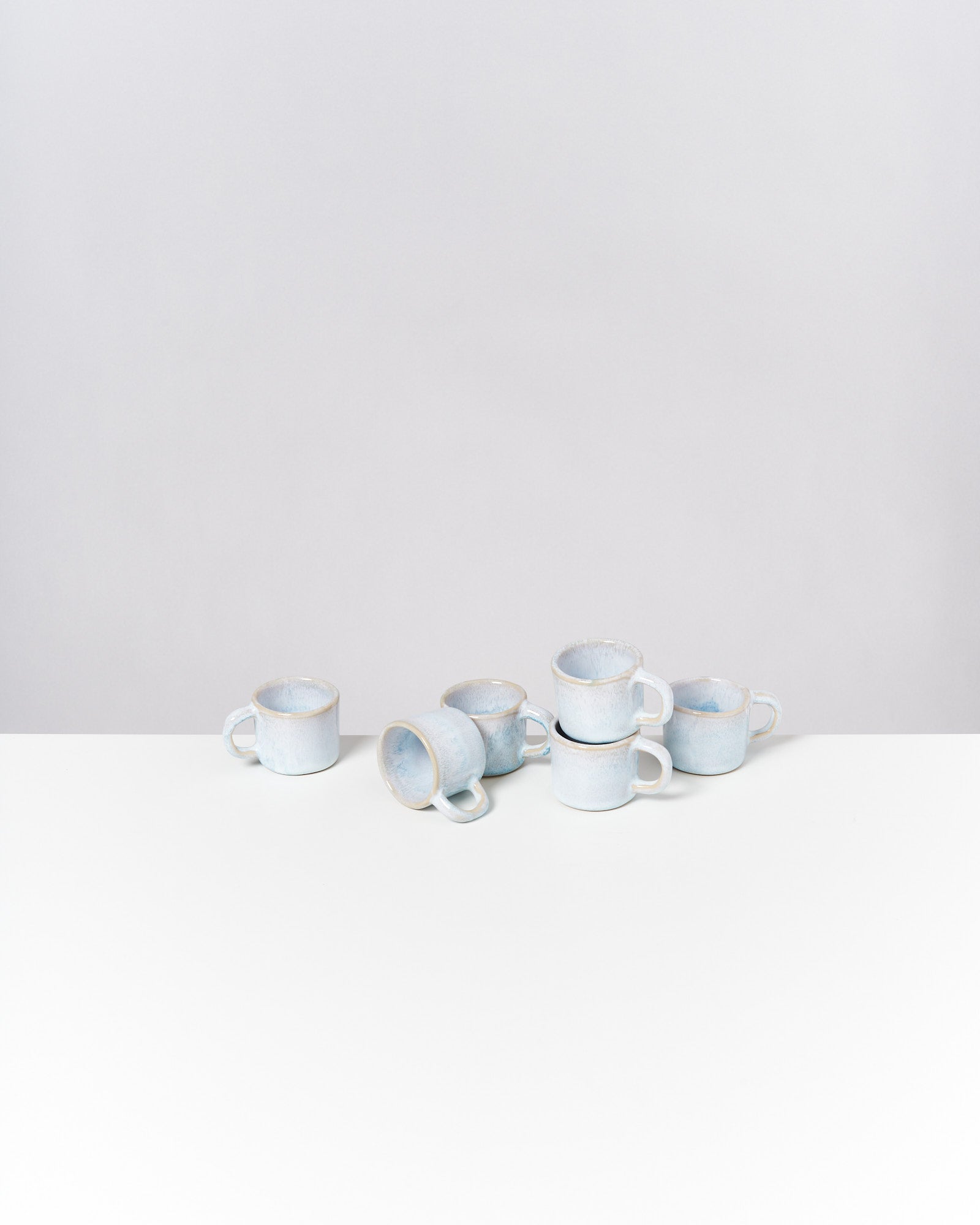 Mehrere stapelbare, hellblaue Tassen stehen und liegen auf einer weißen Oberfläche vor grauem Hintergrund.
