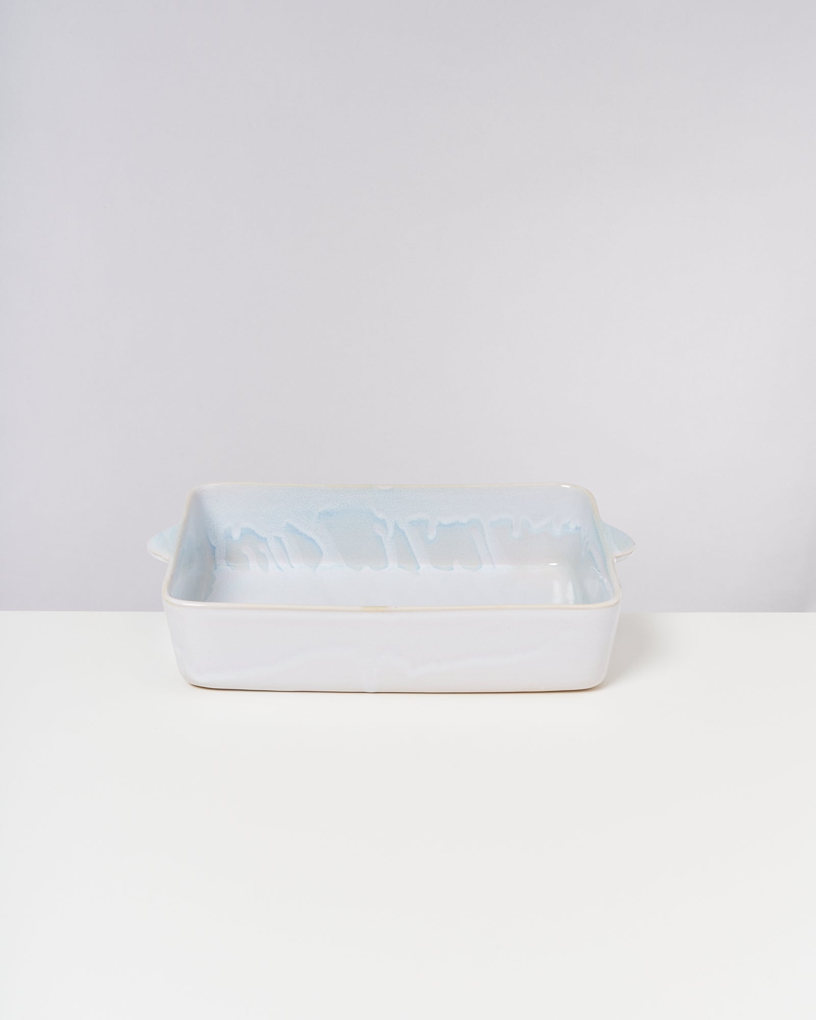 Weiße rechteckige Keramikauflaufform mit seitlichen Griffen auf einer weißen Oberfläche vor einem hellgrauen Hintergrund.