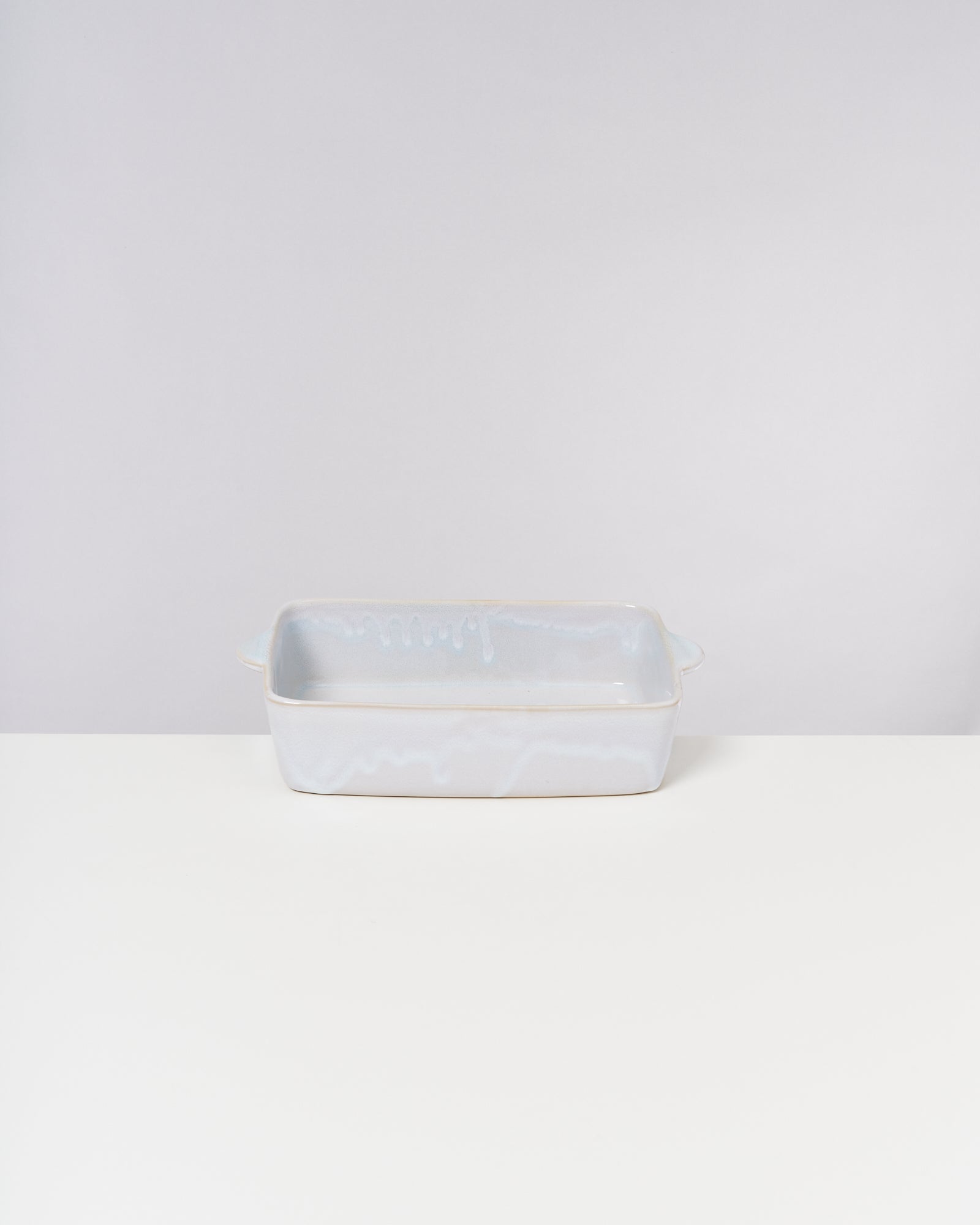 Weiße, rechteckige Keramikauflaufform mit Griffen, auf einem weißen Tisch vor grauem Hintergrund.