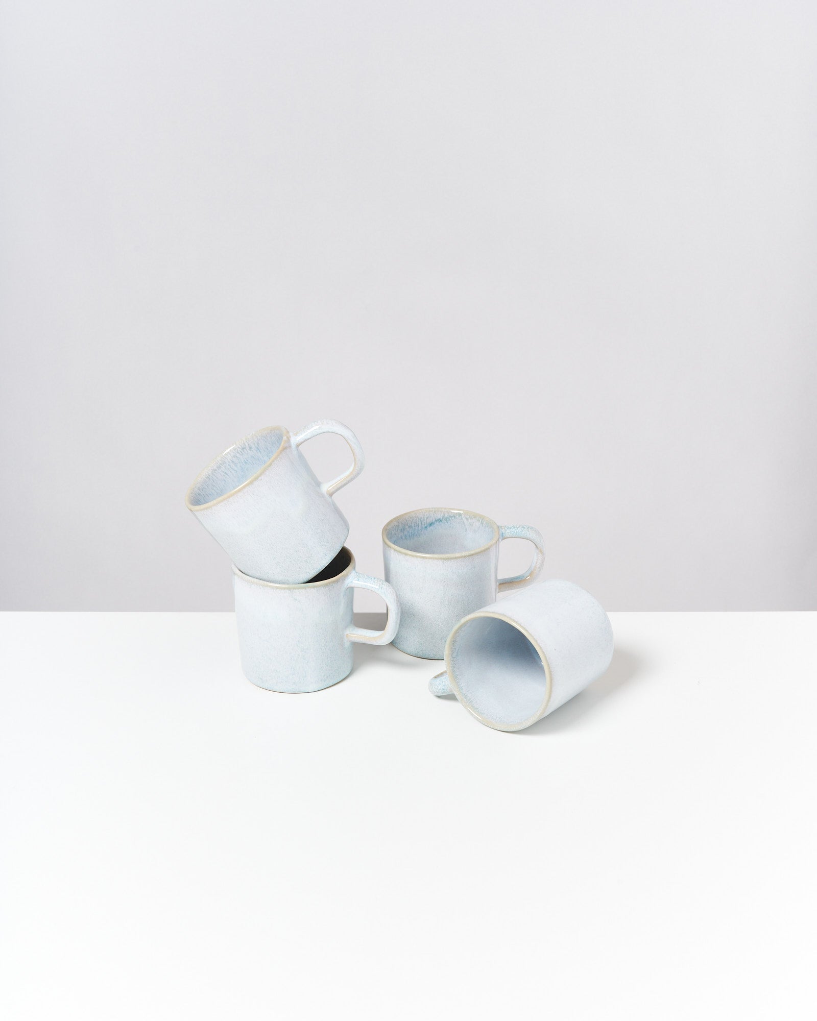 Vier weiße Keramikbecher stapeln sich auf einem weißen Tisch vor einem grauen Hintergrund.