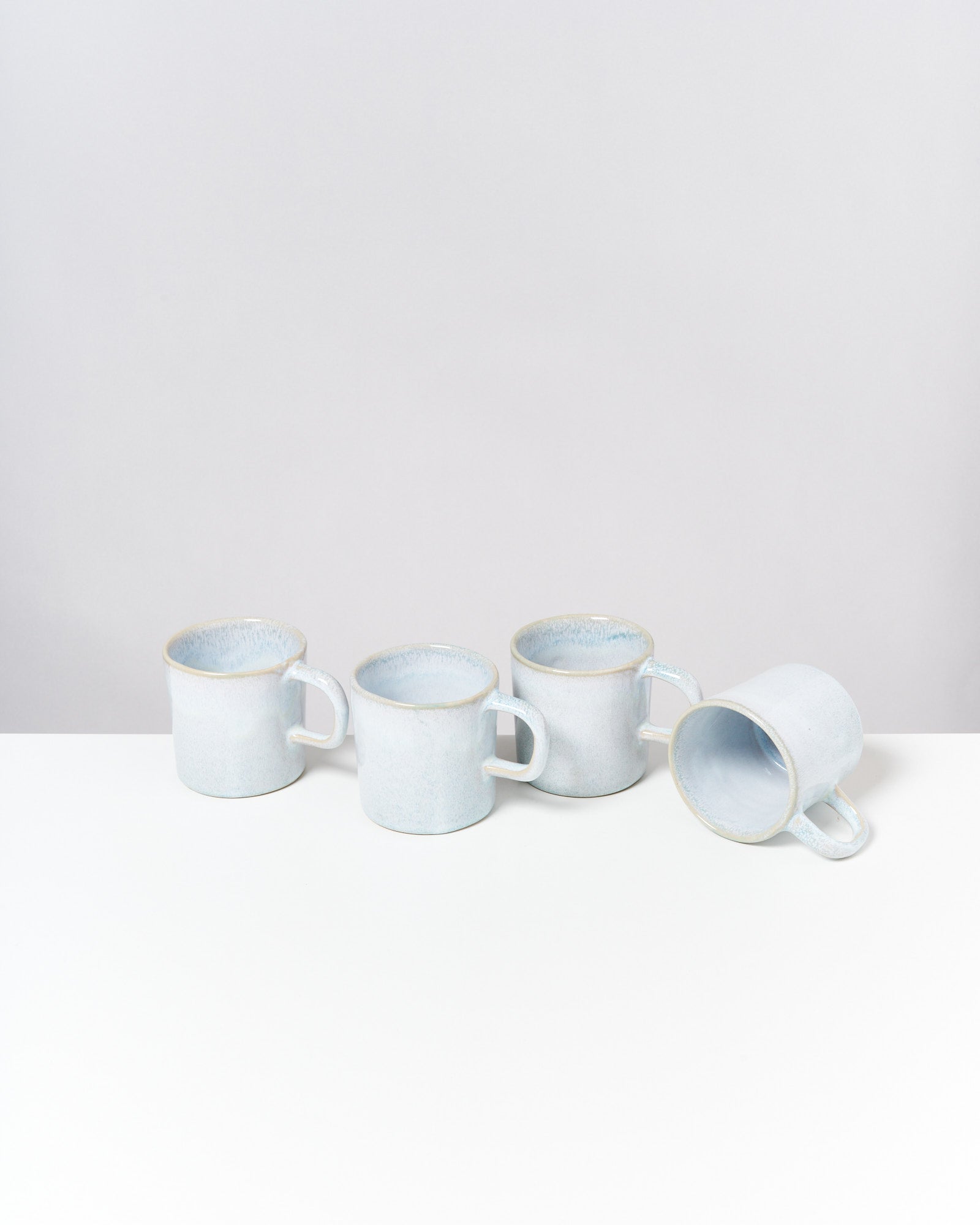 Vier blaue Keramiktassen, drei aufrecht stehend und eine liegend, auf einem weißen Tisch vor grauem Hintergrund.