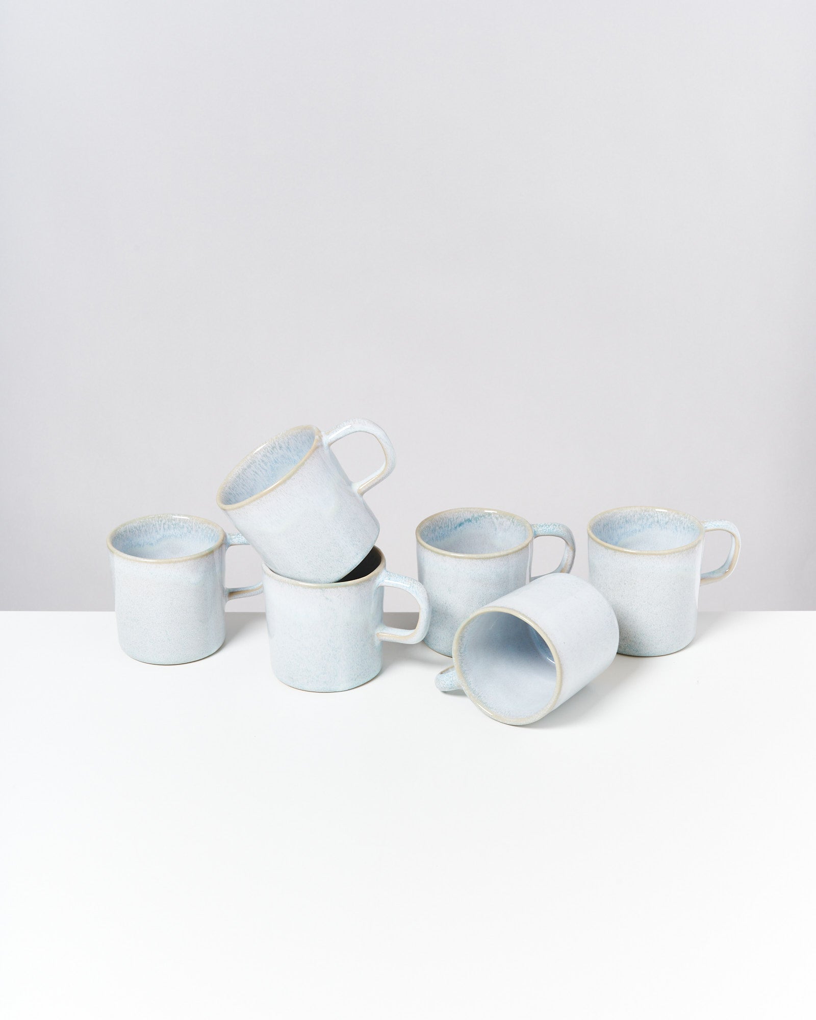 Sechs hellblaue Keramiktassen stehen und liegen in einem Stapel auf einer weißen Oberfläche.