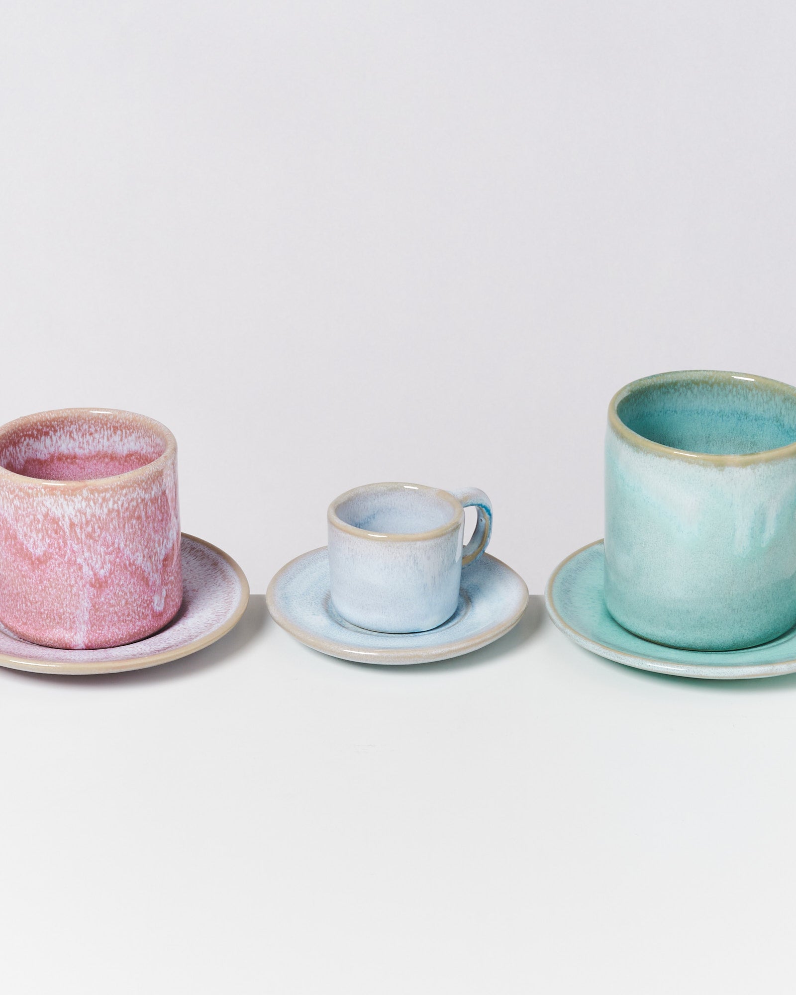 Drei Keramiktassen mit passenden Untertassen in Rosa, Blau und Grün auf einem weißen Hintergrund.