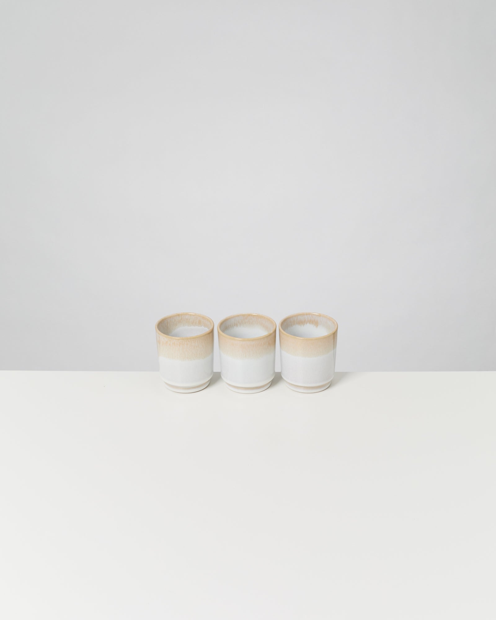 Drei weiße Keramiktassen mit braunem Rand stehen auf einem weißen Tisch vor einem grauen Hintergrund.