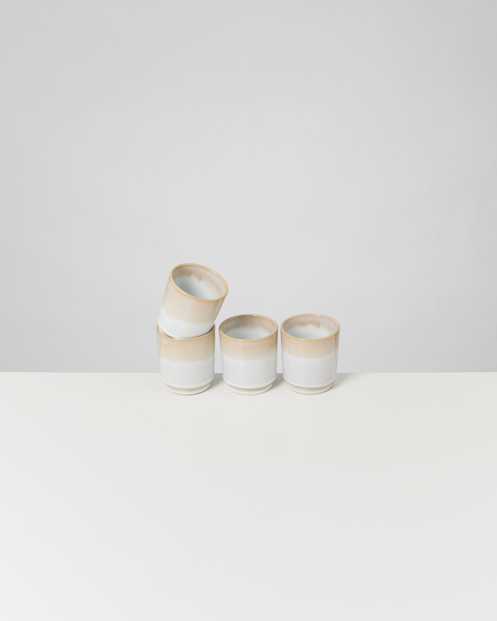 Vier beige und weiße Becher, einer schräg gestapelt, auf einem weißen Tisch vor grauem Hintergrund.
