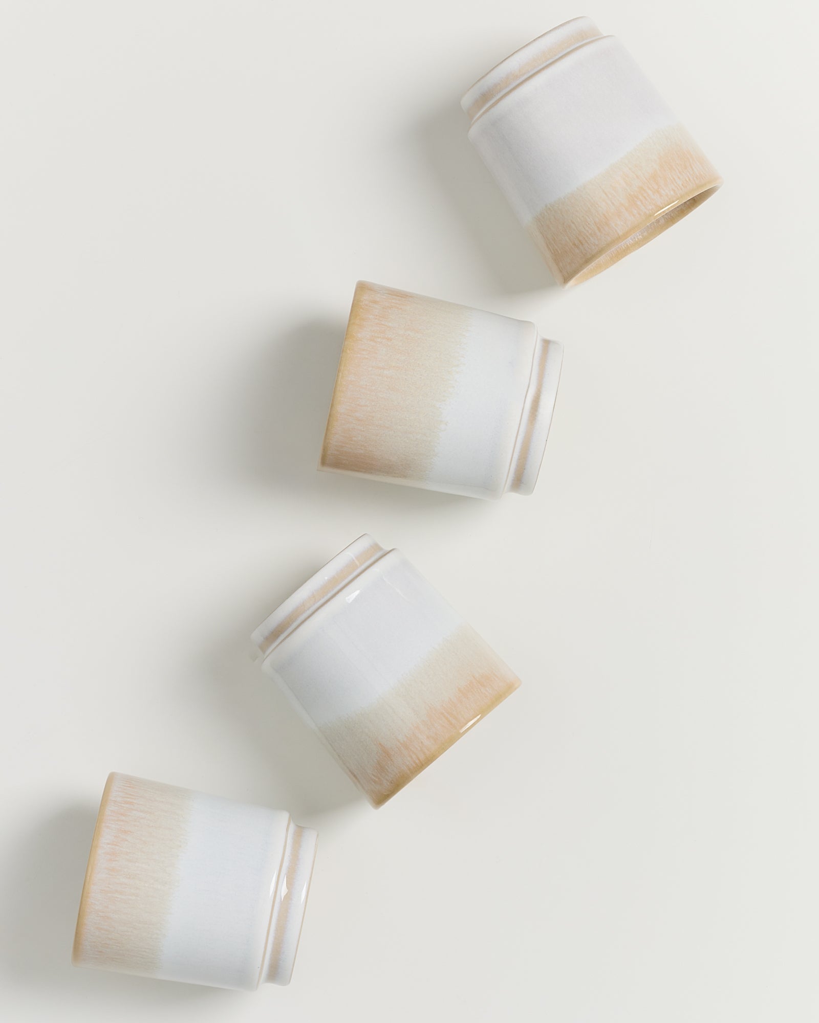 Vier umgekippte, weiß-beige Keramikbecher liegen diagonal auf einer hellen Oberfläche.