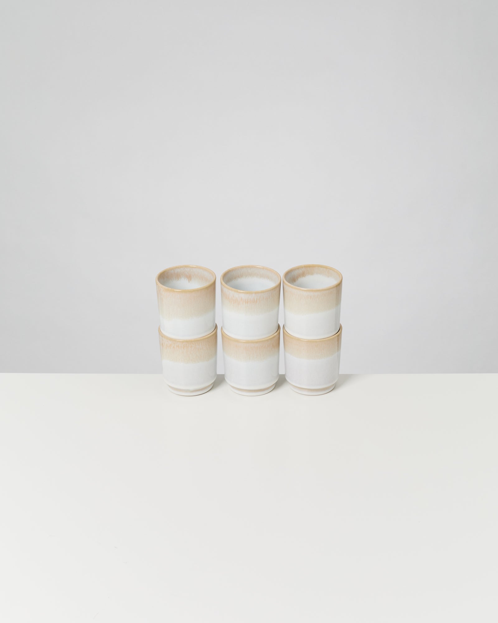 Sechs gestapelte, beige-weiße Keramiktassen auf weißem Untergrund.