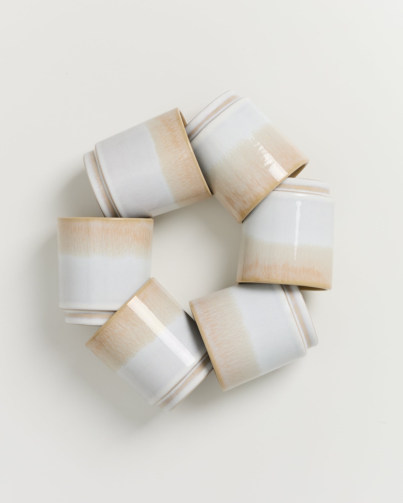 Sechs helle Keramikbecher, die im Kreis auf einer weißen Oberfläche angeordnet sind.