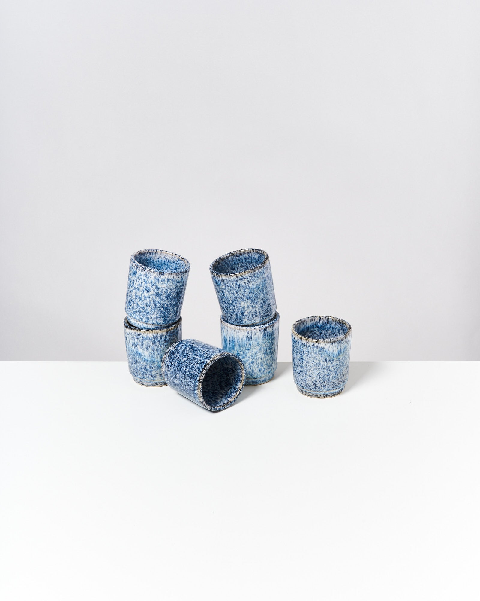 Sechs blaue, gesprenkelte Keramikbecher stapeln sich auf einem weißen Tisch vor einem weißen Hintergrund.