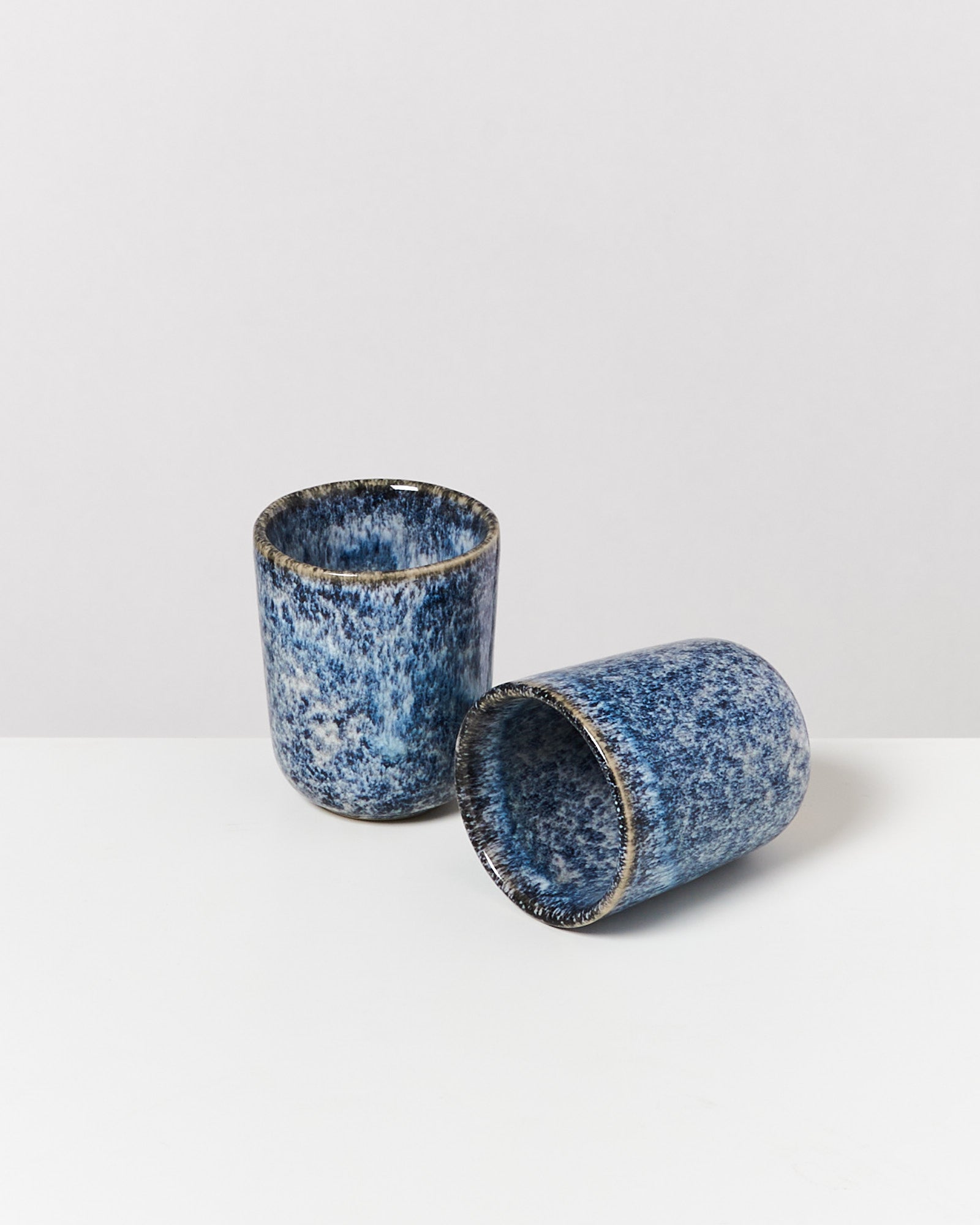 Zwei blau gemusterte Keramiktassen, eine stehend und die andere liegend, auf einem weißen Untergrund.