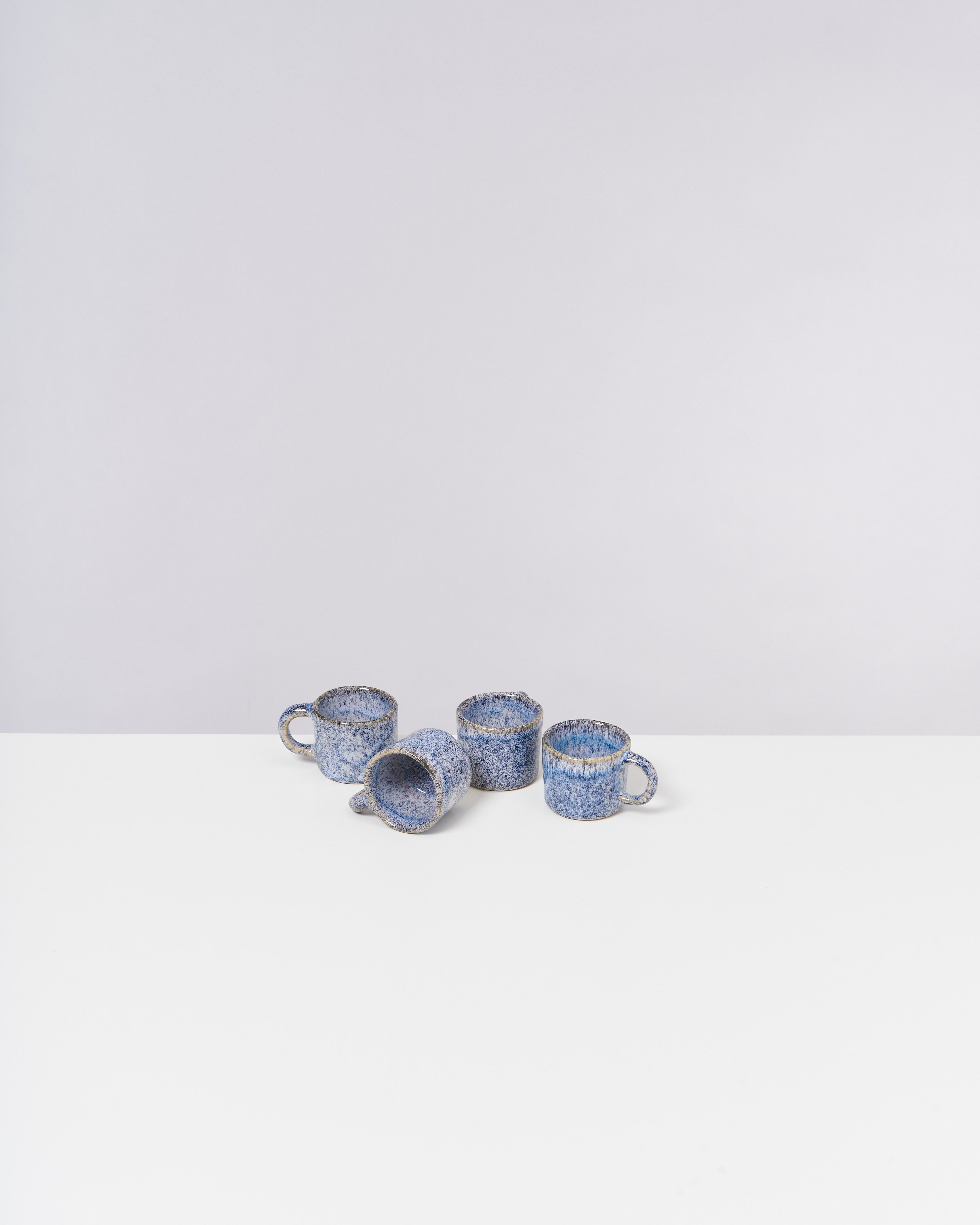 Vier blaue, gesprenkelte Tassen stehen auf einer weißen Oberfläche, eine davon liegt seitlich.