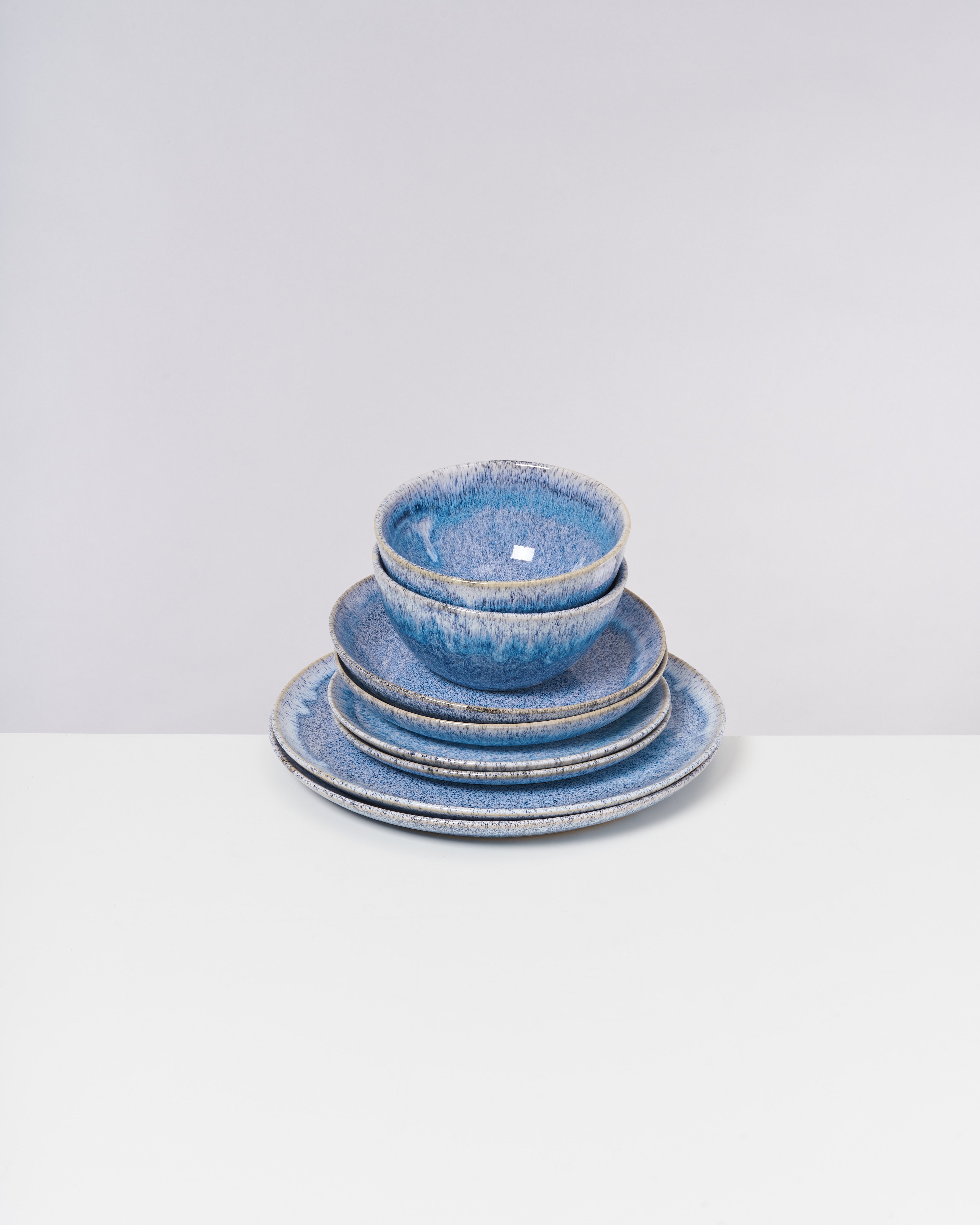 Ein blaues Keramikgeschirr-Set aus zwei Schüsseln und drei Tellern auf einem weißen Tisch vor grauem Hintergrund.