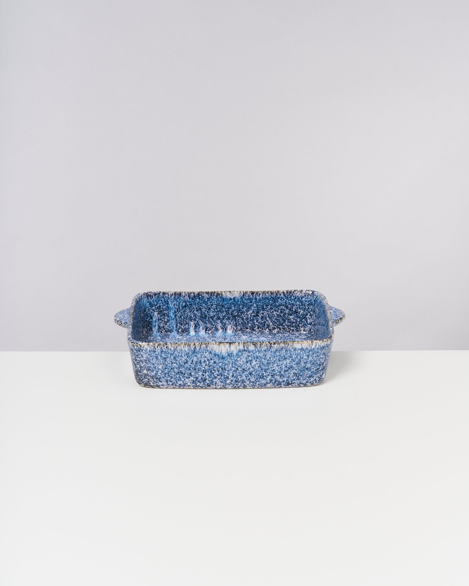 Blau gesprenkelte Keramik-Auflaufform mit Griffen auf einem weißen Tisch vor grauem Hintergrund.