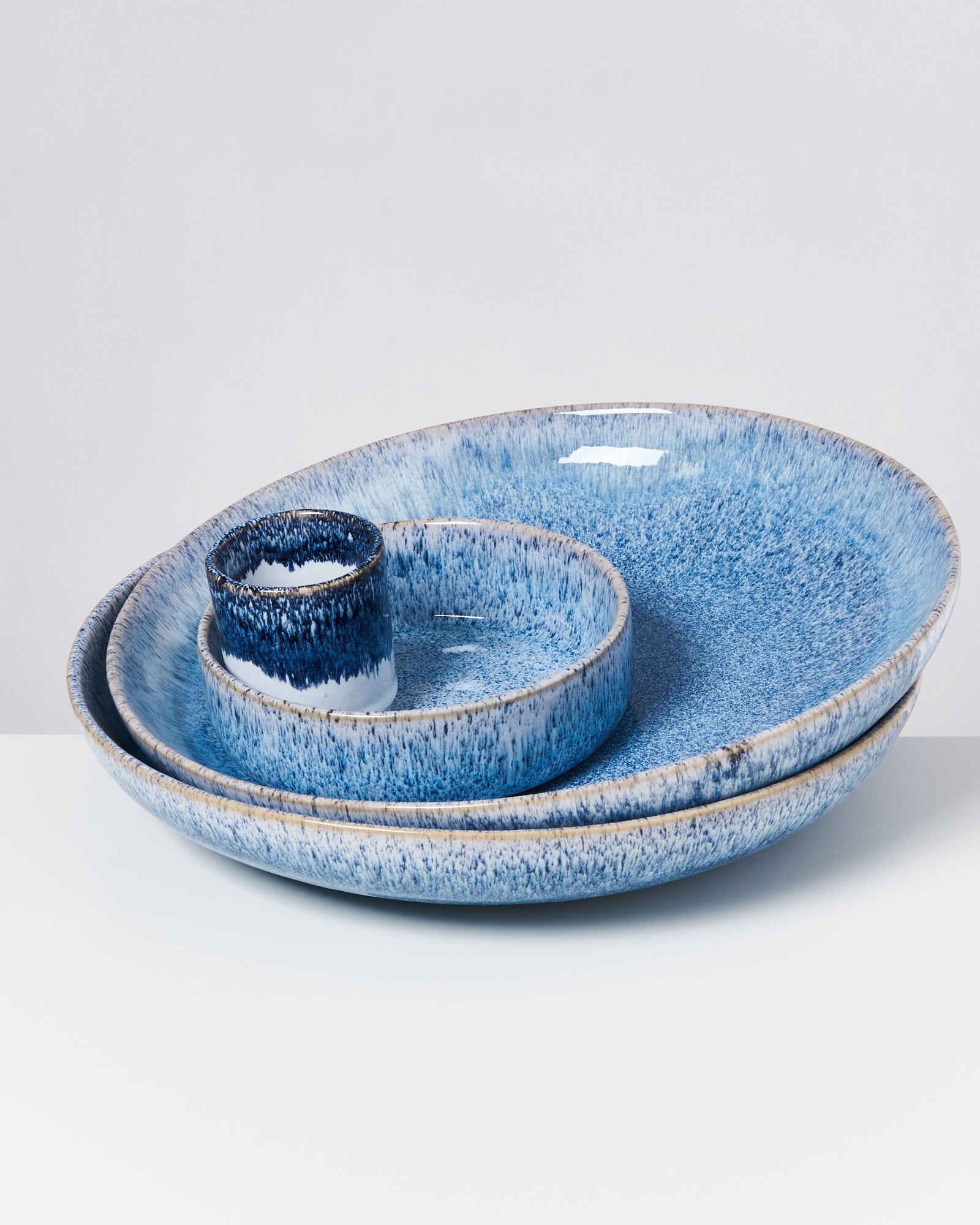 Drei blaue Keramikteller und ein Becher mit einem gesprenkelten Muster, gestapelt auf einem weißen Hintergrund.
