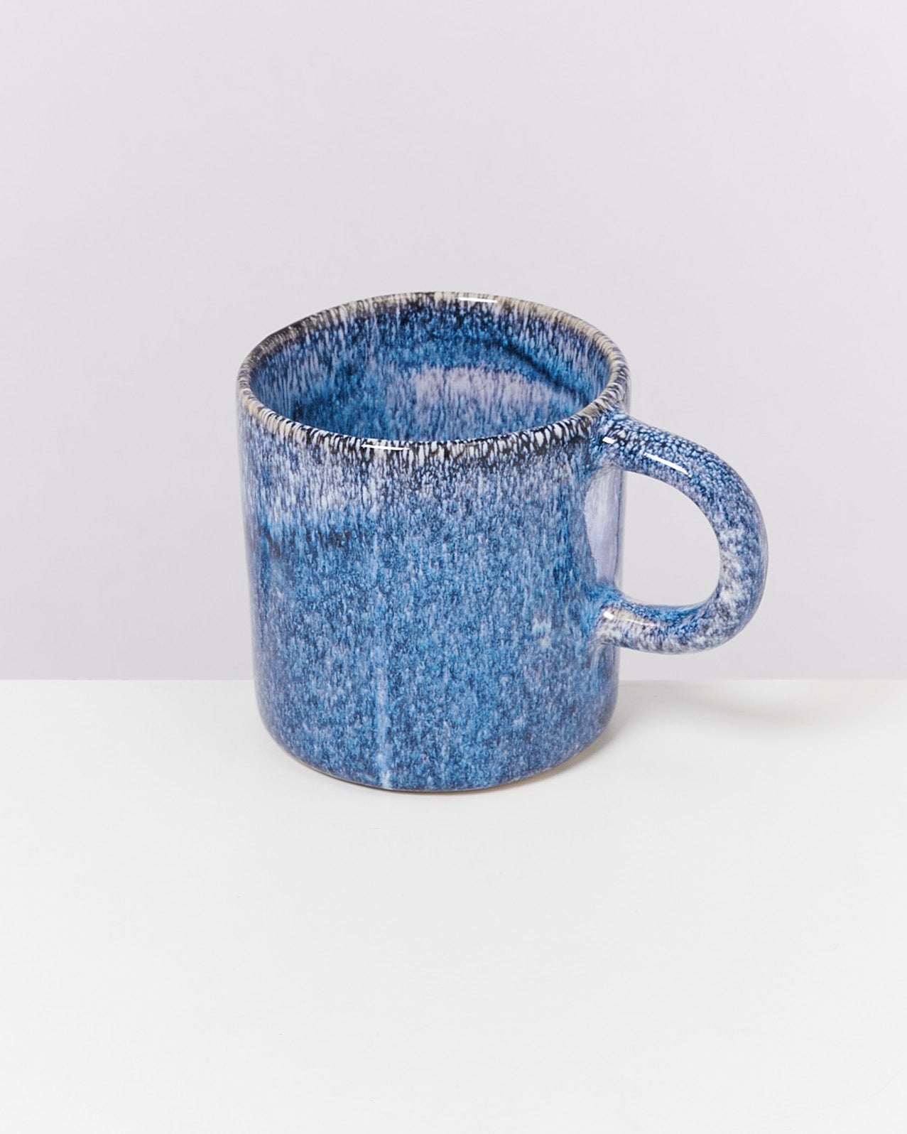 Blauer Keramikbecher mit gräulicher Maserung auf einem weißen Tisch vor einem hellen Hintergrund.
