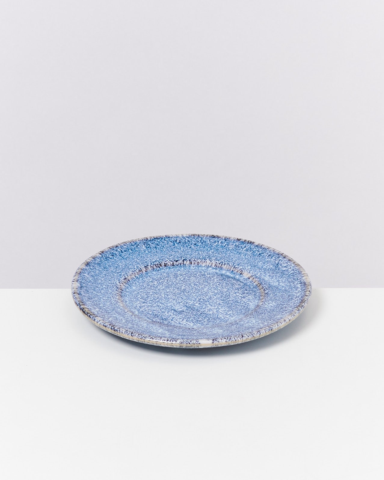 Ein blauer Keramikteller mit gesprenkeltem Muster auf einer weißen Oberfläche vor grauem Hintergrund.