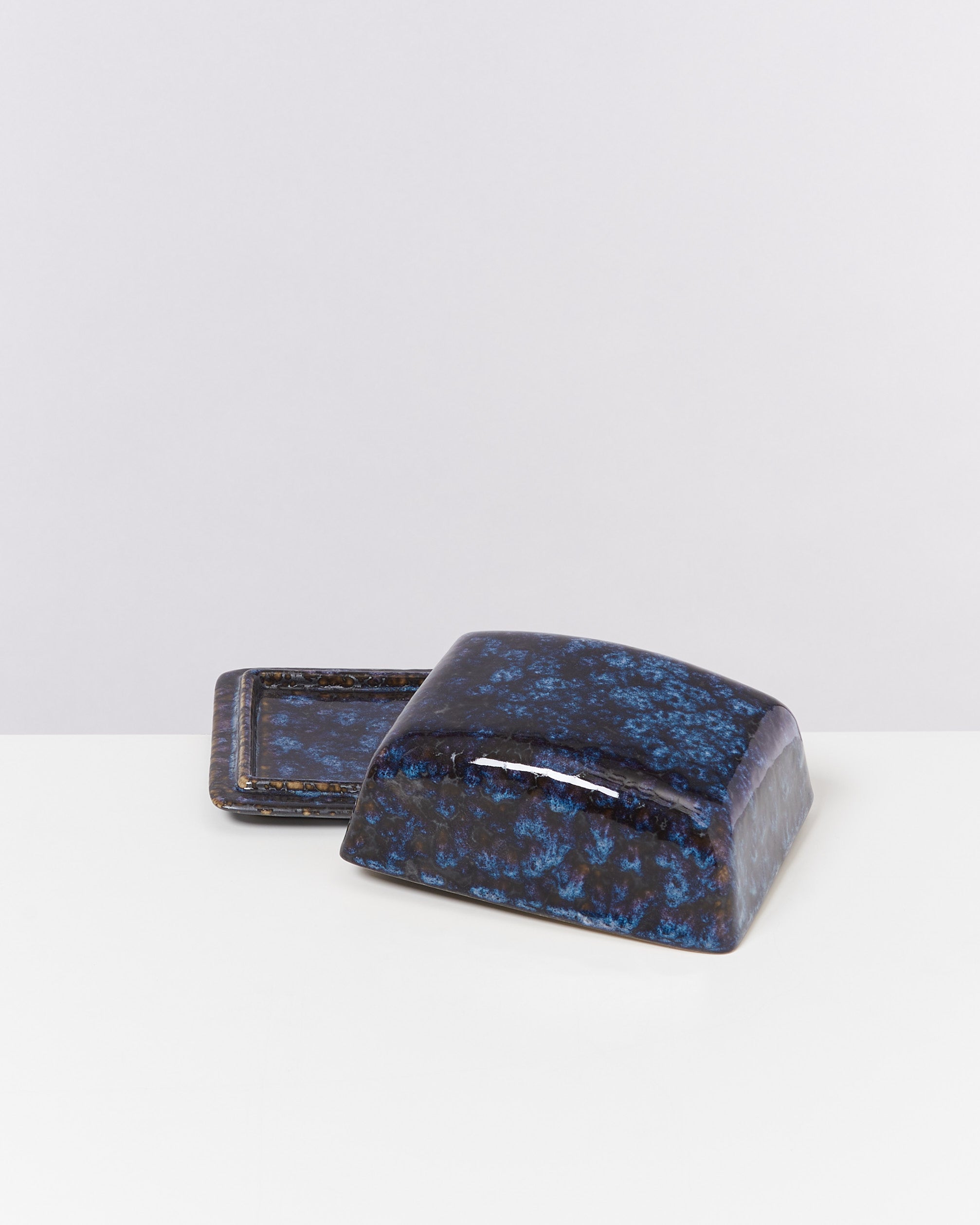 Ein rechteckiger Deckel mit blauer und schwarzer Glasur liegt auf einem passenden Unterteil auf einem weißen Hintergrund.