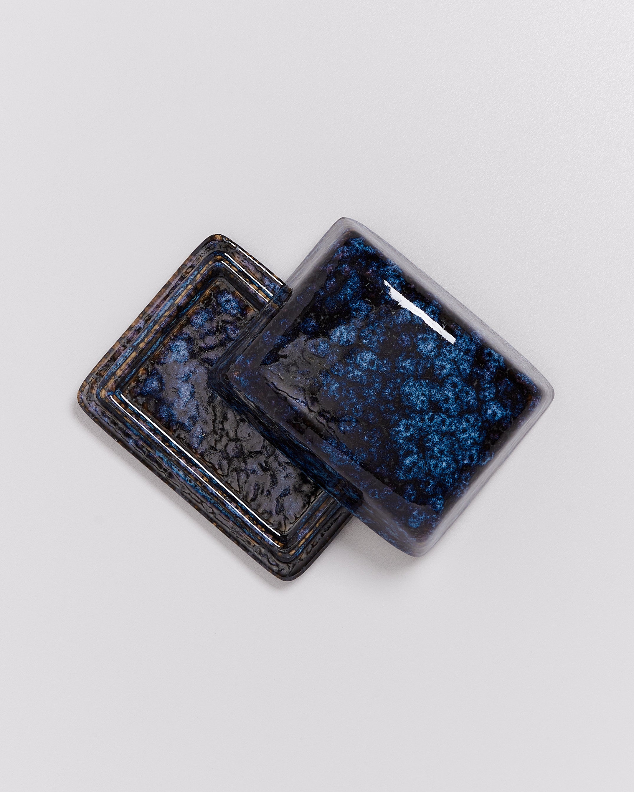 Zwei übereinandergelegte quadratische Keramikschalen mit blauer, gesprenkelter Glasur auf einer grauen Oberfläche.