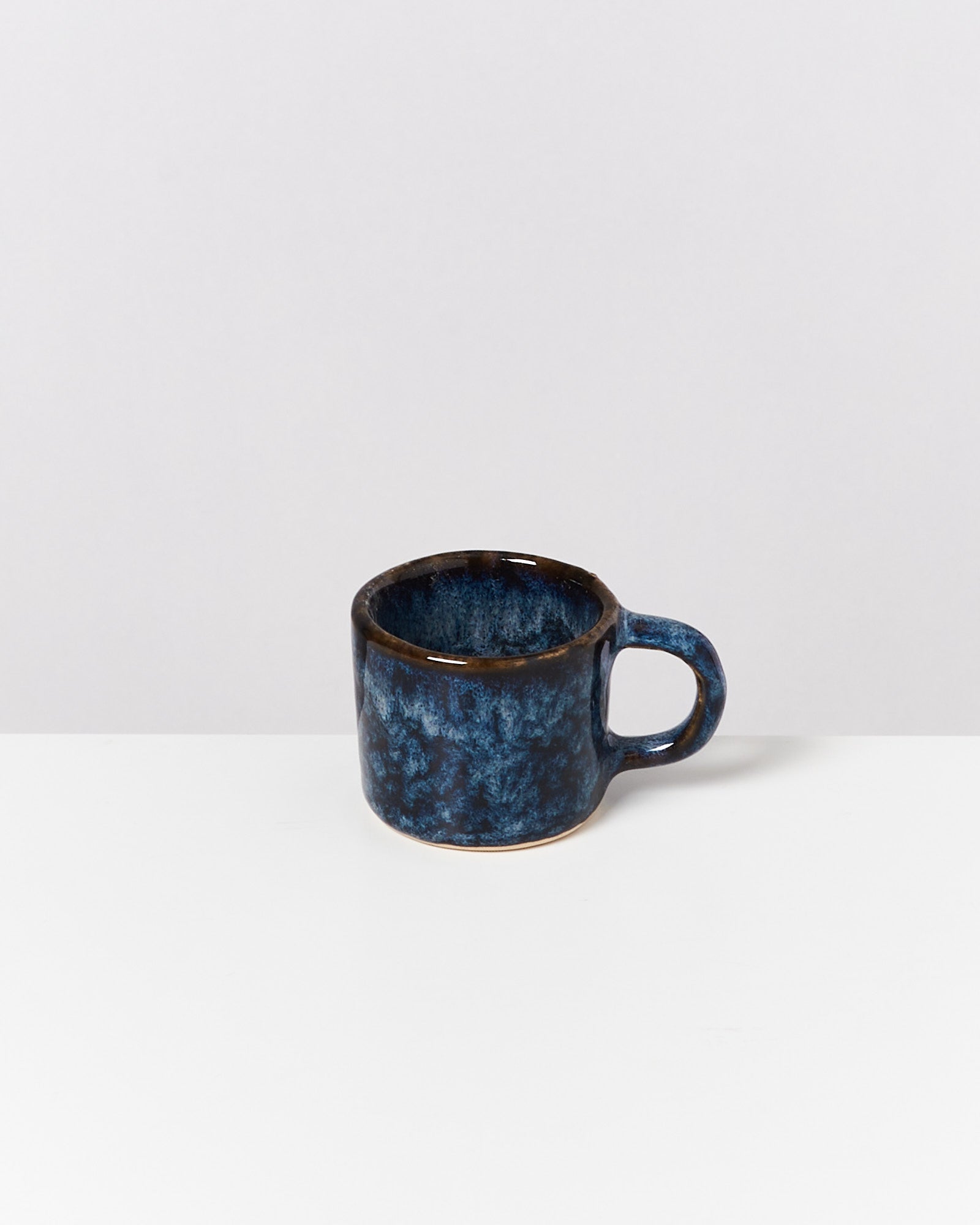 Blaue Keramiktasse mit Henkel steht auf einem weißen Untergrund vor einem grauen Hintergrund.