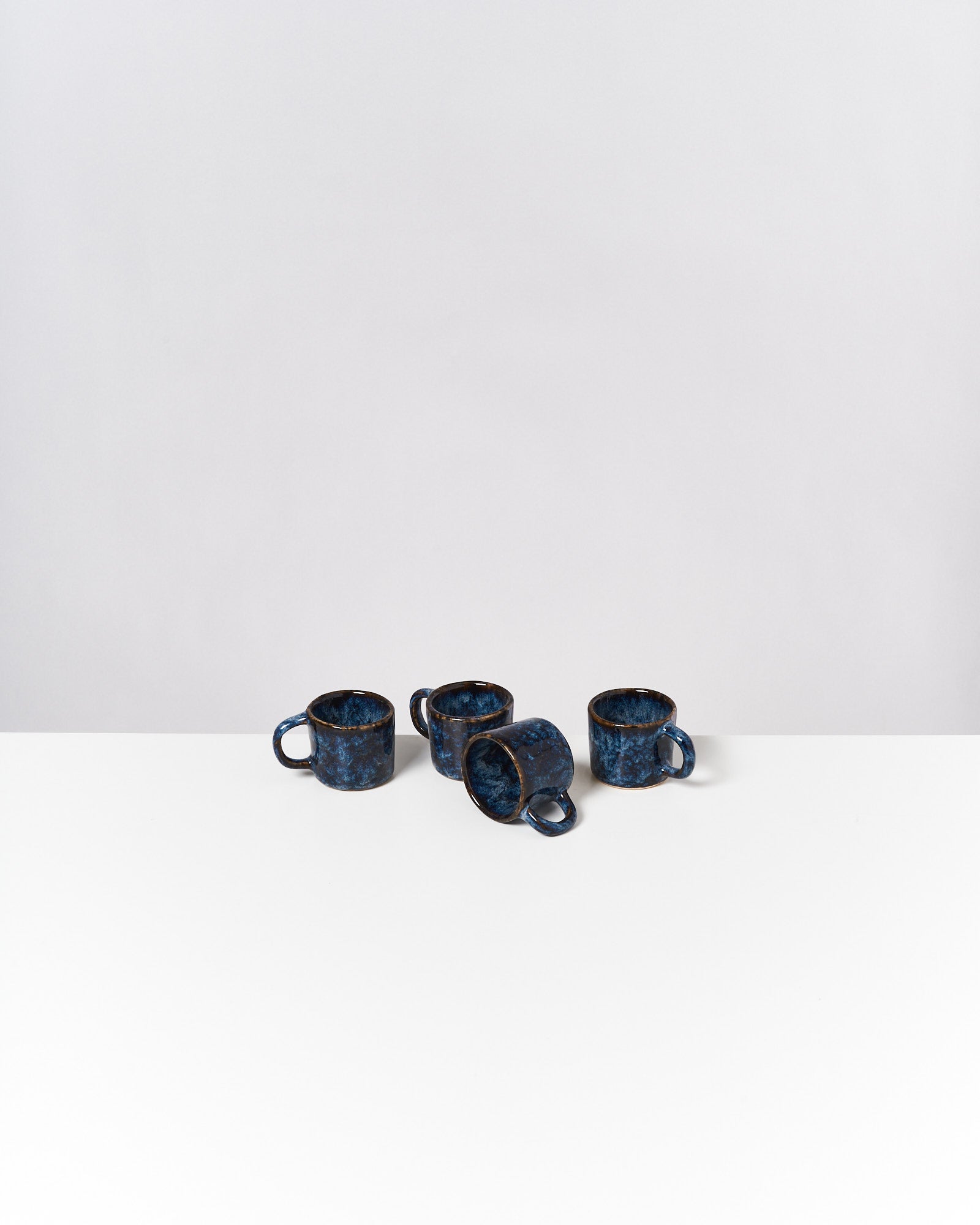 Vier blaue Keramiktassen, eine liegend, auf einem weißen Tisch vor grauem Hintergrund.