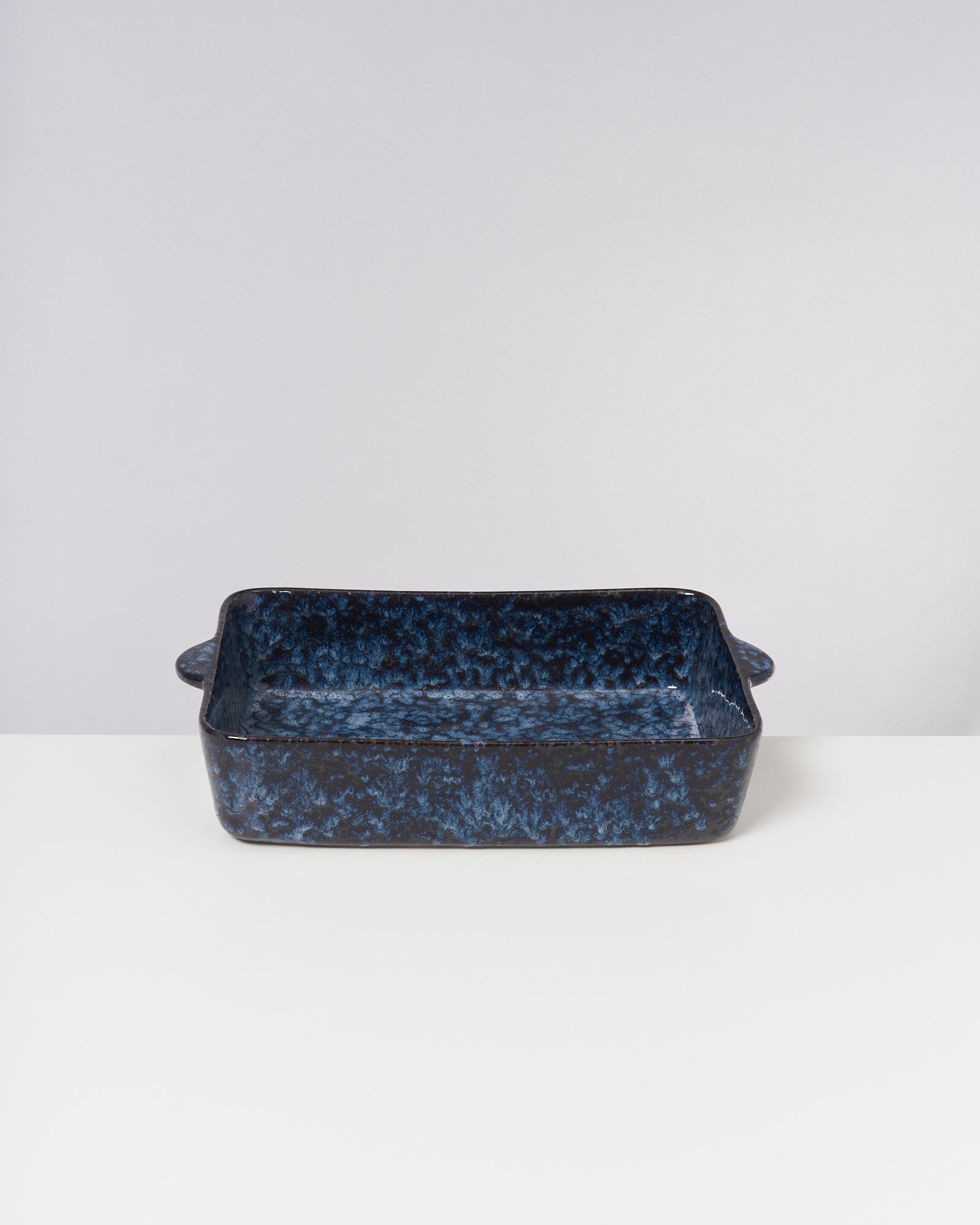 Blaue rechteckige Keramikbackform mit Griffen auf einem weißen Tisch vor einem neutralen Hintergrund.