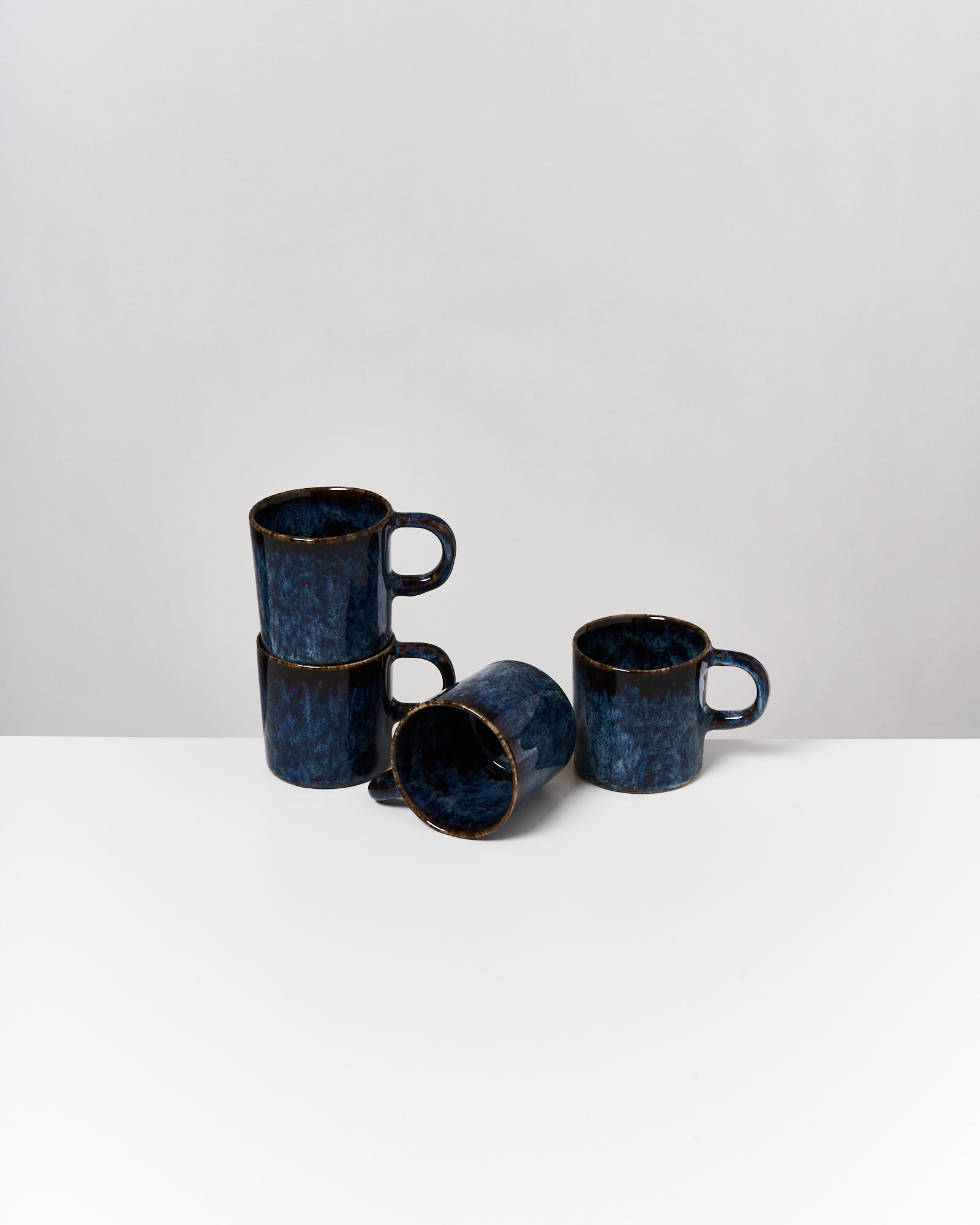 Vier blaue Tassen, von denen drei gestapelt und eine liegend, auf einer weißen Oberfläche vor neutralem Hintergrund.