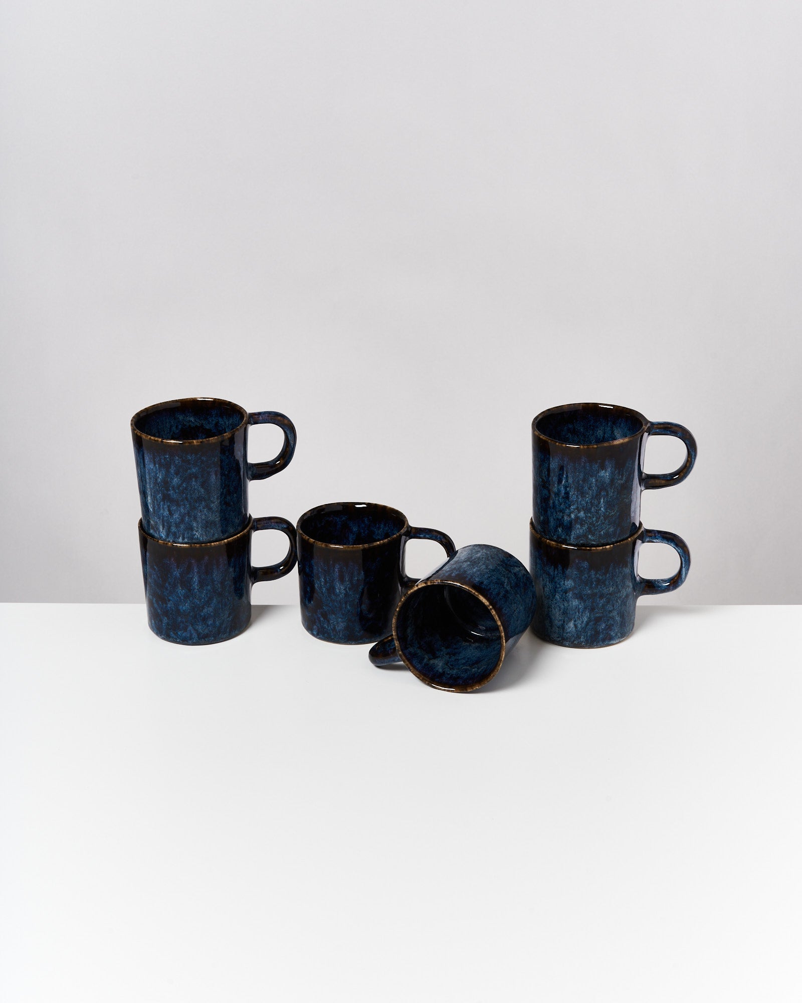 Sechs blaue Tassen aus Keramik, vier gestapelt und zwei liegen auf der Seite, auf einer weißen Oberfläche.