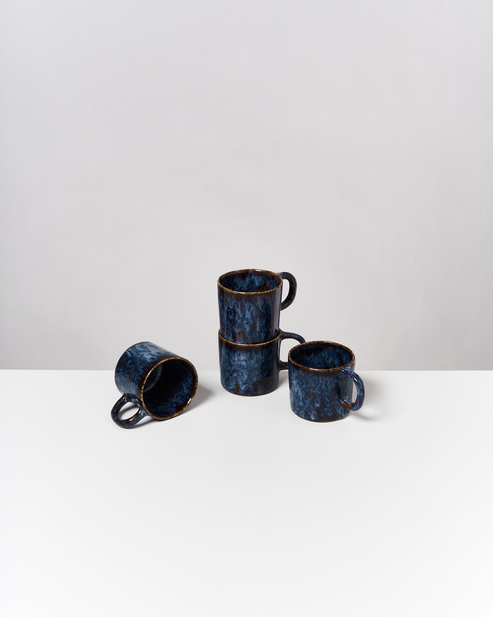 Drei gestapelte blaue Tassen und eine umgefallene Tasse auf einem weißen Tisch vor neutralem Hintergrund.