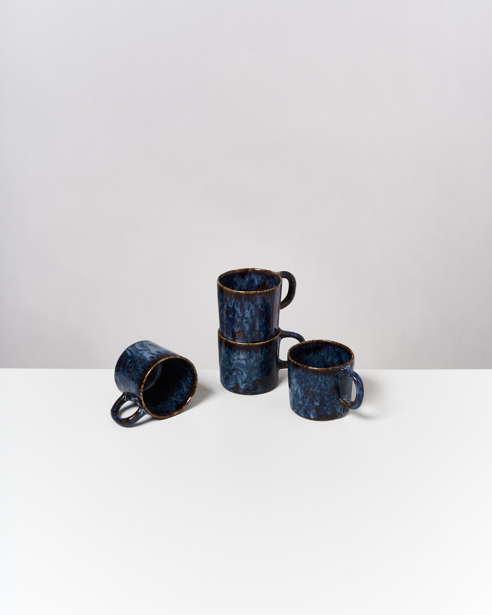 Vier blaue Keramiktassen, drei gestapelt und eine liegend, auf einem weißen Tisch gegen eine graue Wand.