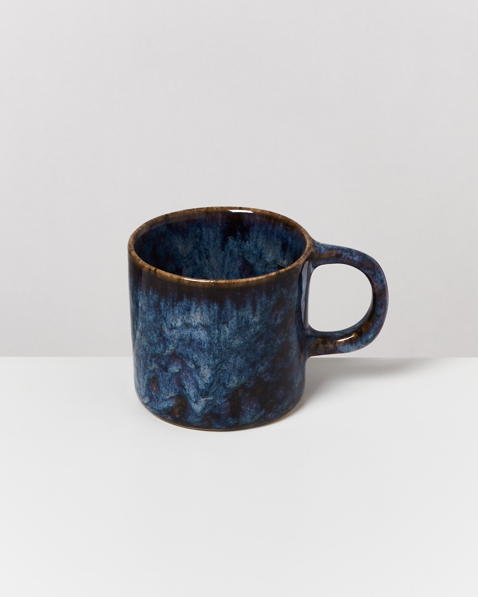 Dunkelblaue Keramiktasse mit einem robusten Henkel steht auf einer weißen Fläche vor einem grauen Hintergrund.