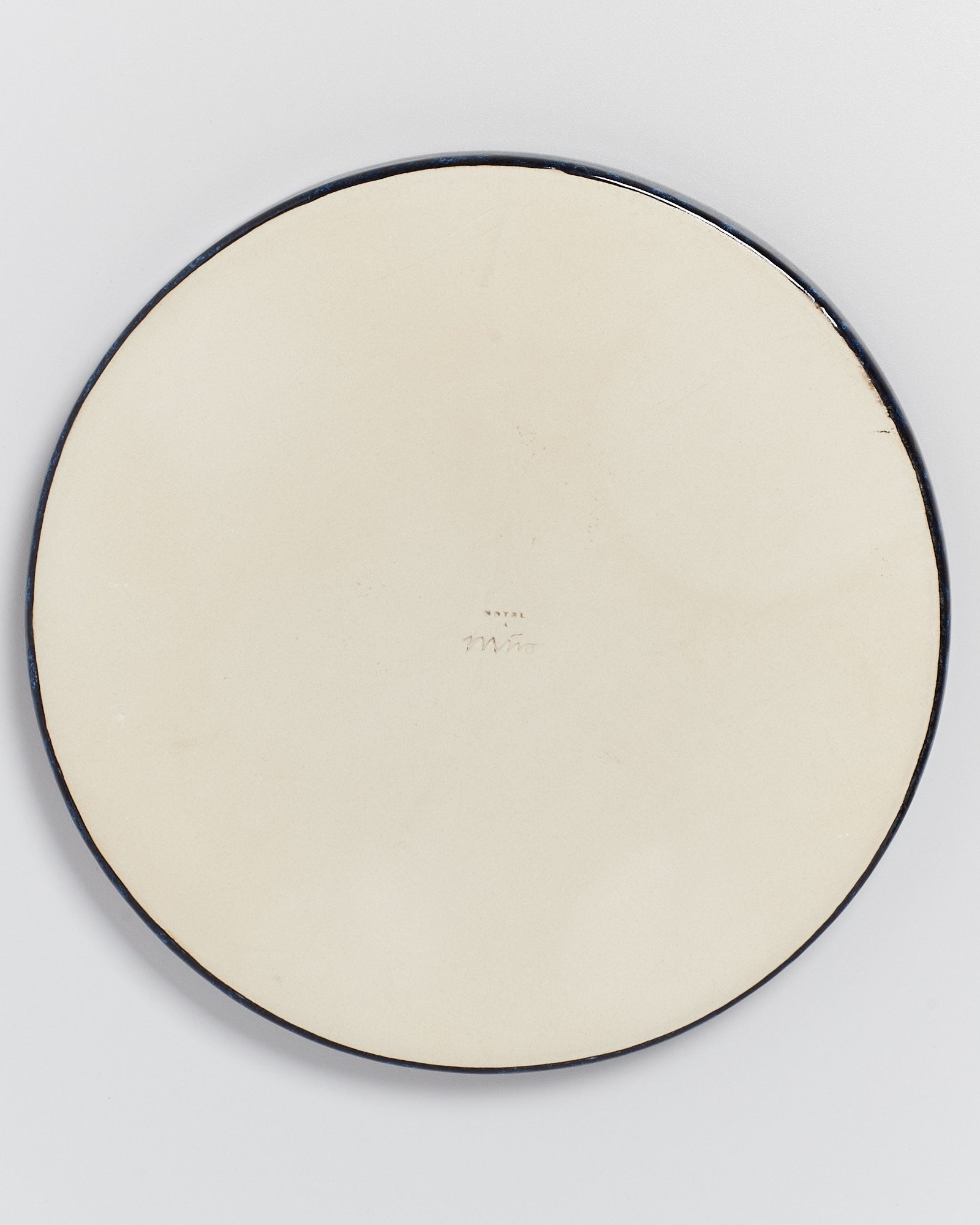 Rückseite eines runden, beigen Tellers mit schwarzem Rand und einer handschriftlichen Signatur in der Mitte.