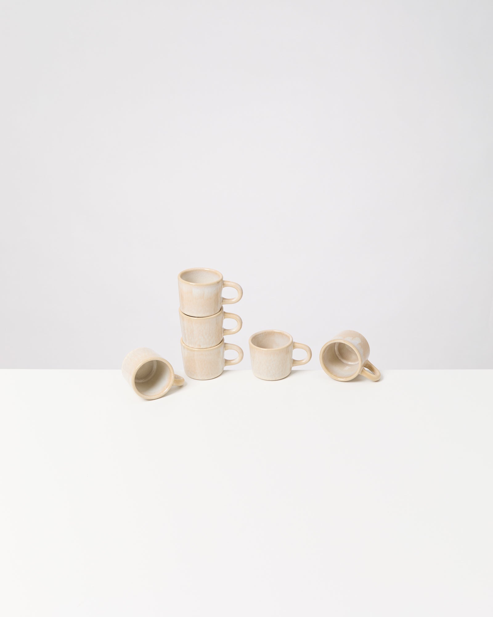 Fünf gestapelte beige Tassen und zwei liegende Tassen auf einem weißen Tisch vor hellem Hintergrund.