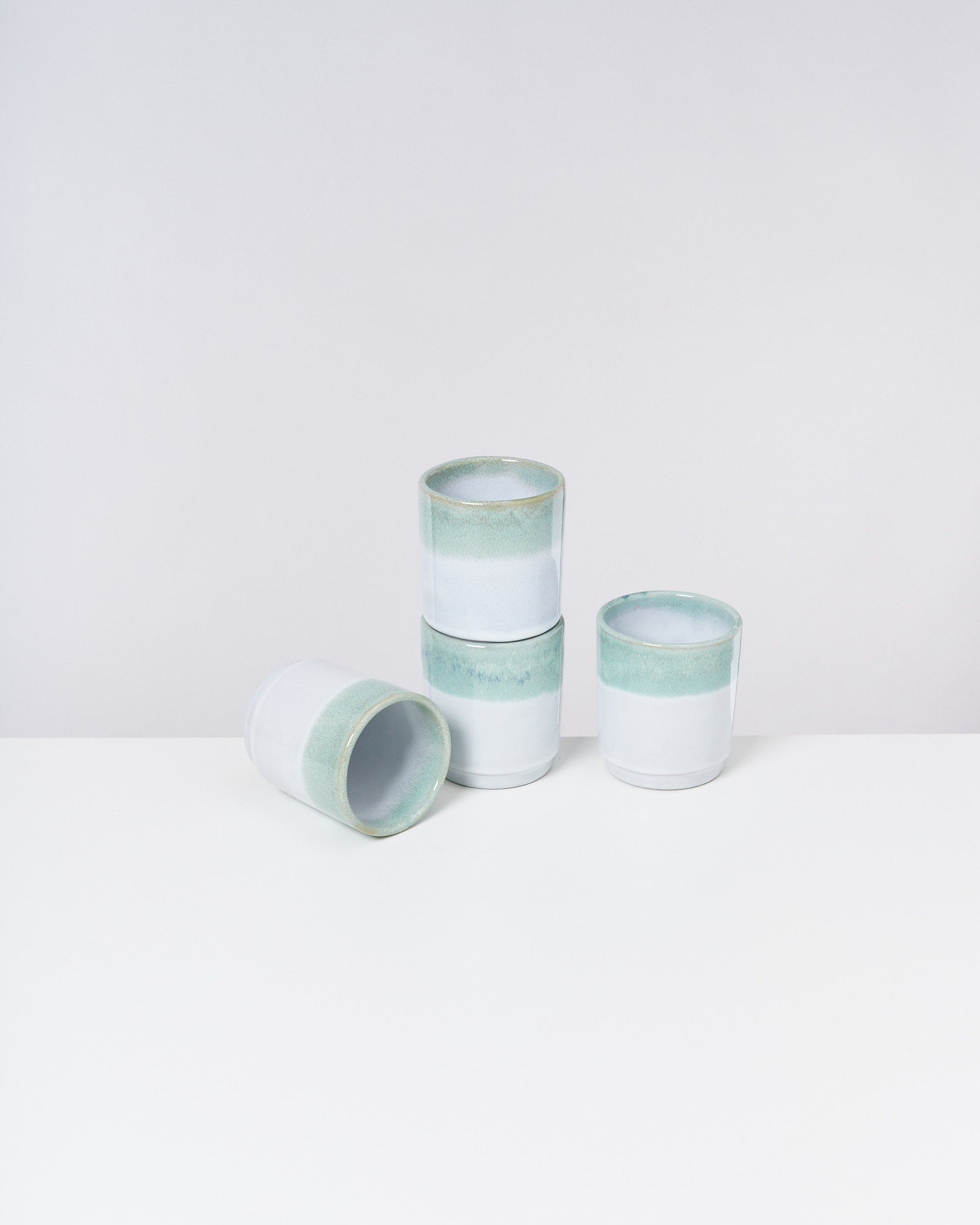Drei stapelbare Keramikbecher in Weiß und Grün mit einem umgekippten Becher daneben auf einem weißen Tisch.