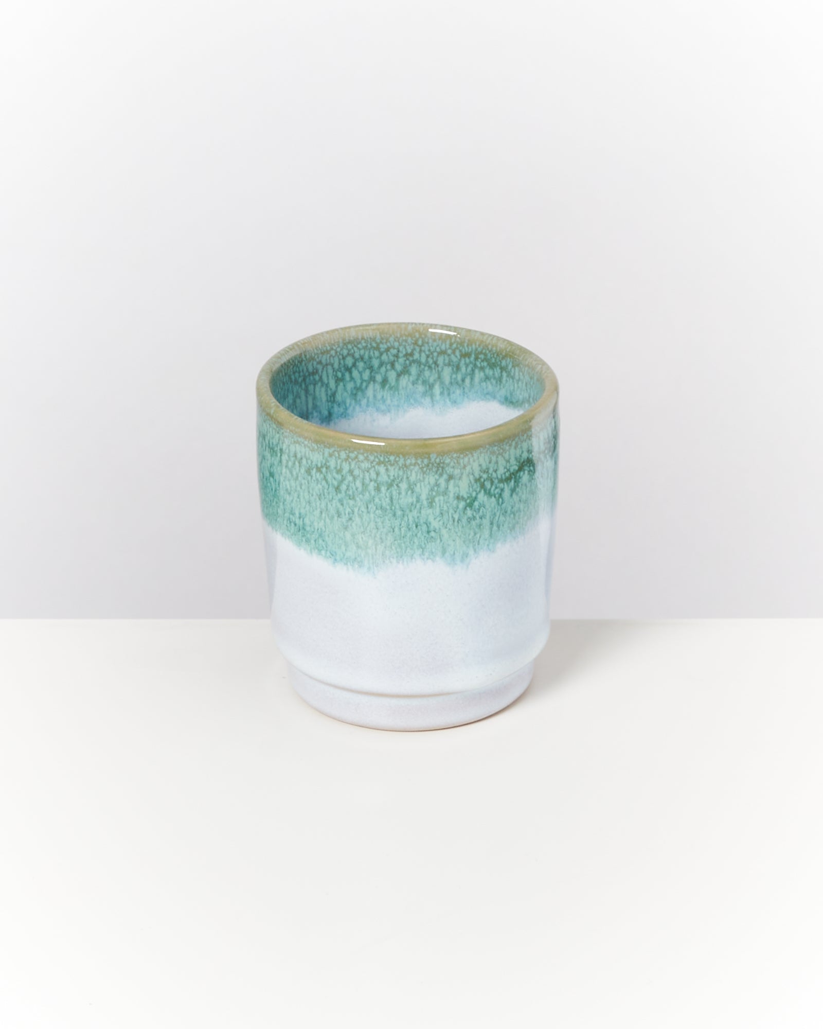 Keramikbecher mit graugrüner Glasur am oberen Rand steht auf einem weißen Hintergrund.