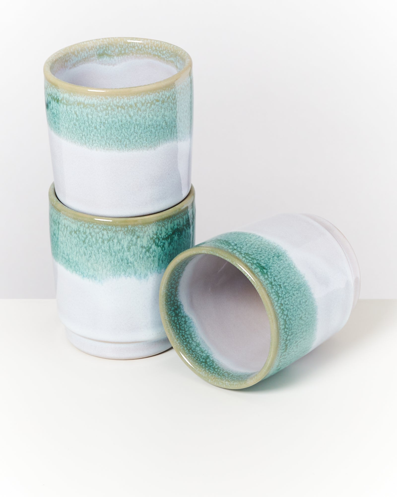 Drei gestapelte Keramikbecher mit weiß-grüner Glasur auf einer weißen Oberfläche.