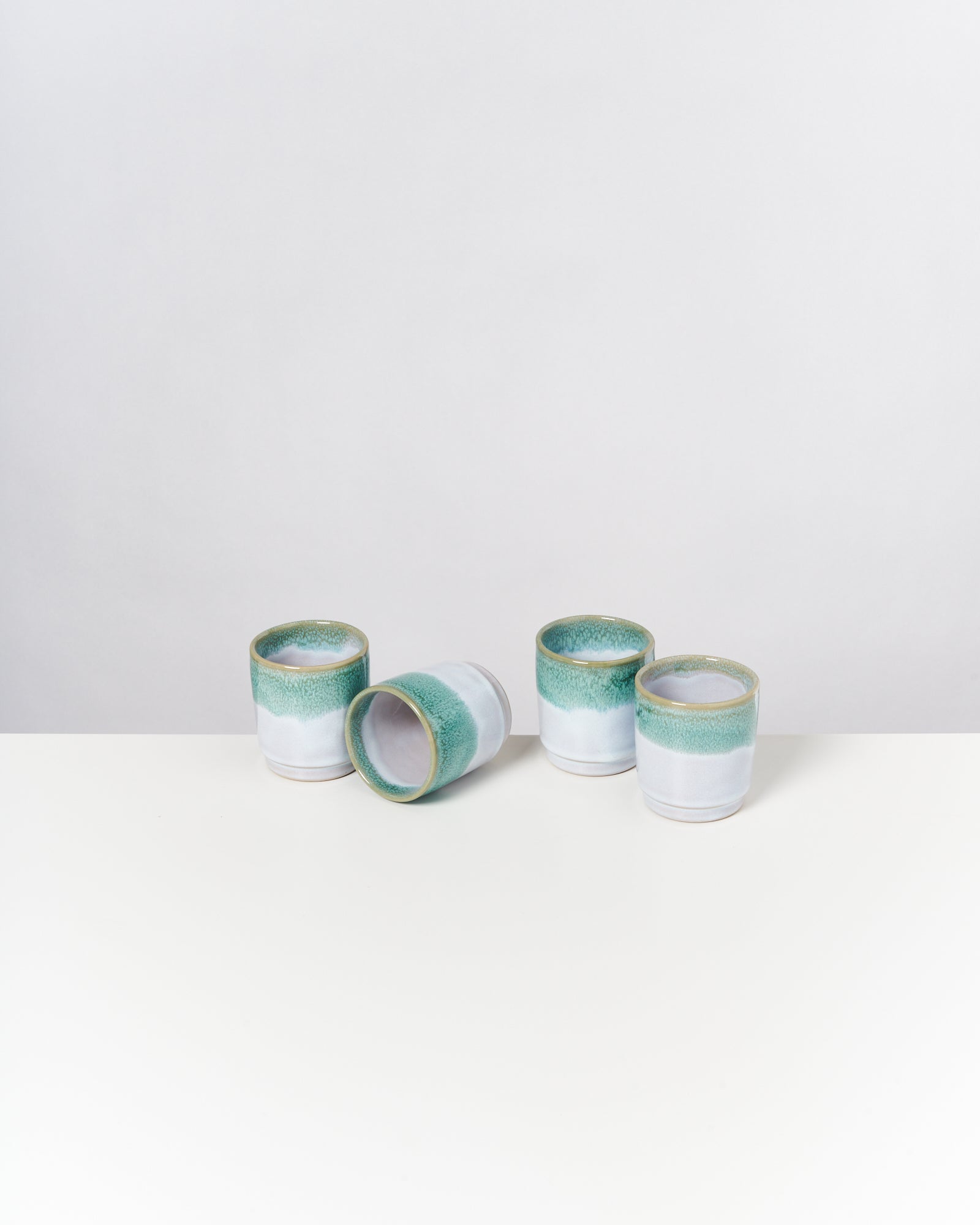 Vier Teetassen mit grüner Glasur auf weißem Hintergrund, eine liegt umgekippt.