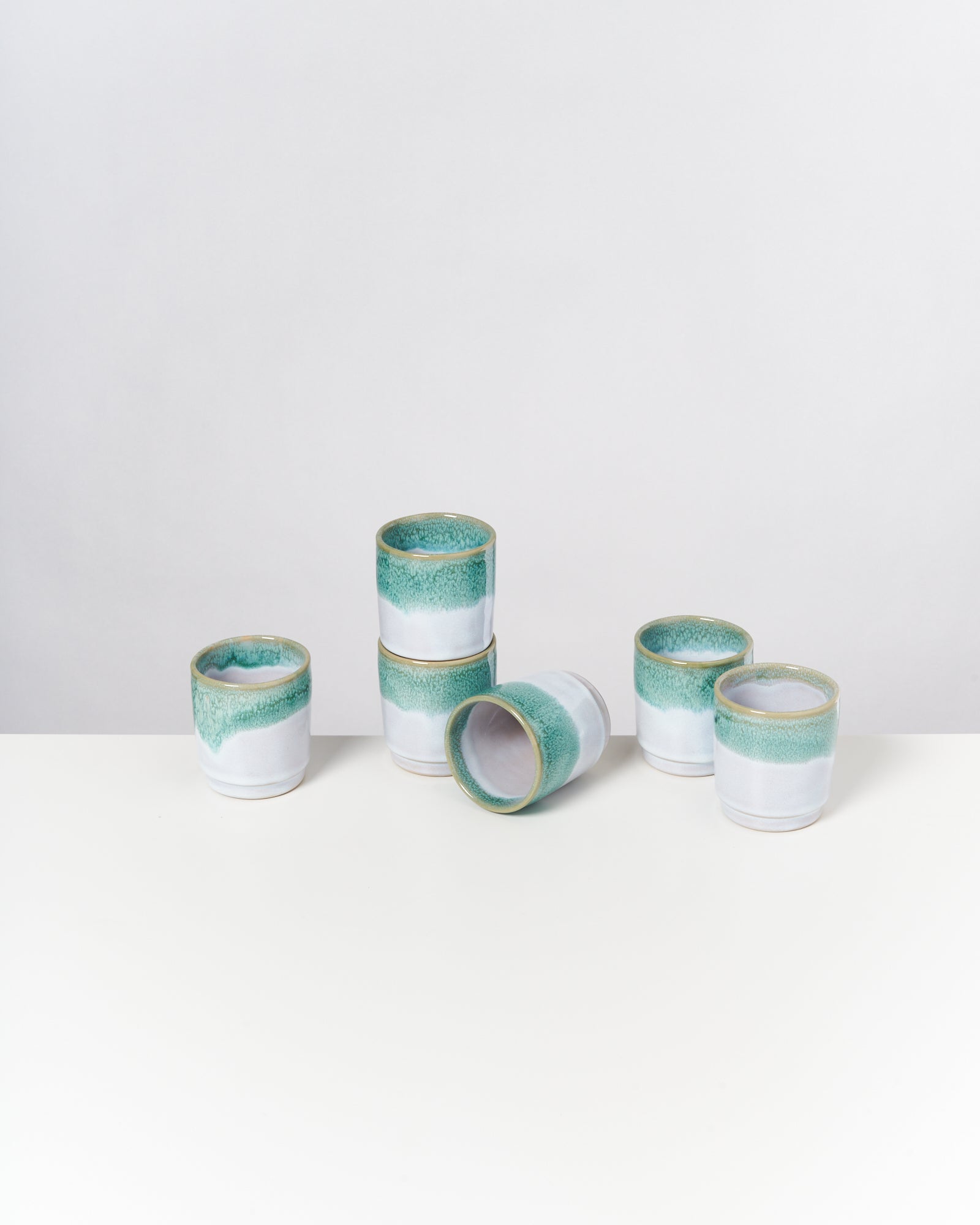 Fünf keramische Becher mit grün-weißer Glasur auf einem weißen Tisch, einer liegt seitlich.